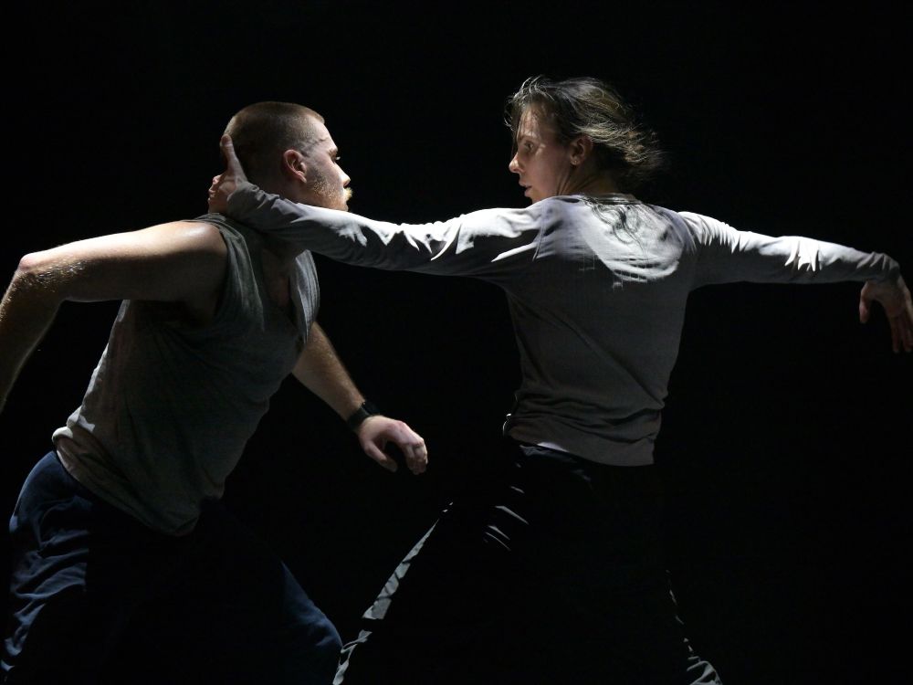 Two dancers stand mid-stride on a stage against a black background. On the left, Justin Rapaport has short strawberry blond hair and a beard. He is wearing a white tank top and dark blue trousers. To the right, Alexis Fletcher is reaching out to Rapaport with a hand on the back of his neck. She has dark hair and her arms are outstretched. She is wearing a white long-sleeved shirt and dark trousers.