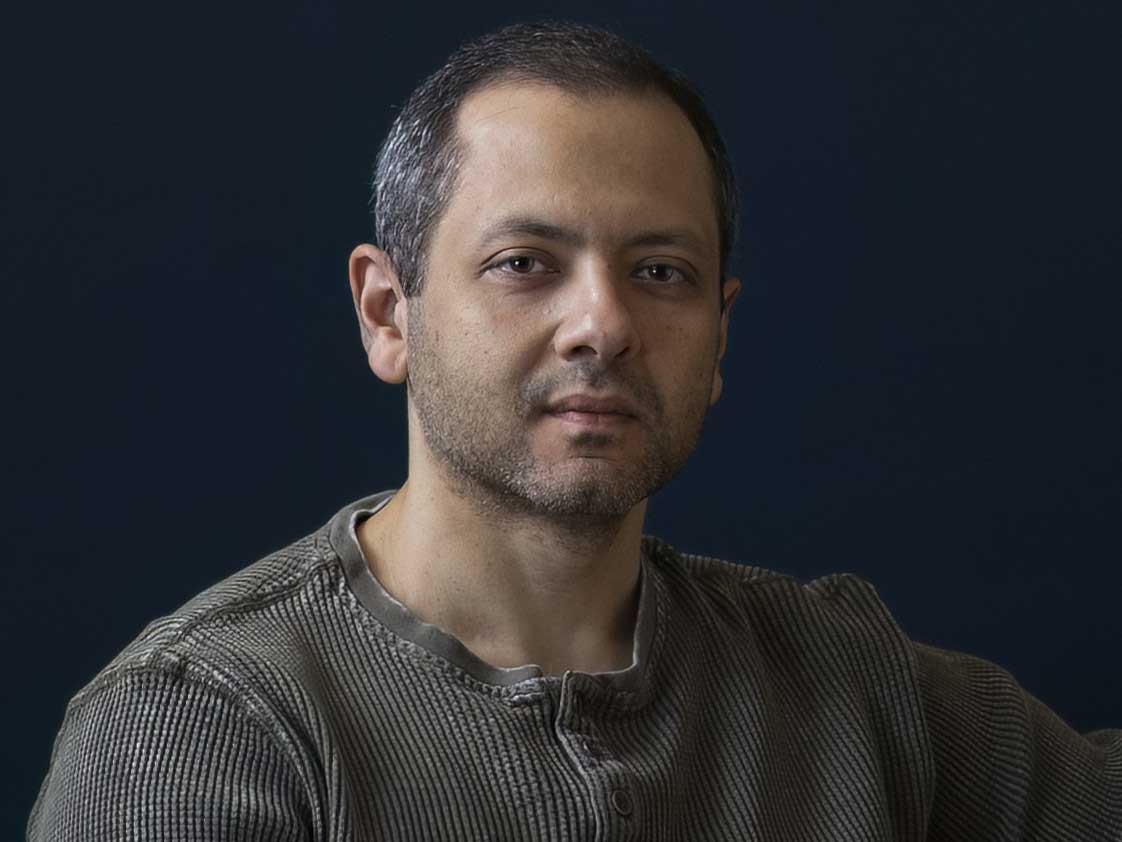 Omar El Akkad has short dark greying hair and is wearing an olive-coloured Henley. He is looking directly at the camera and is seated against a black background.