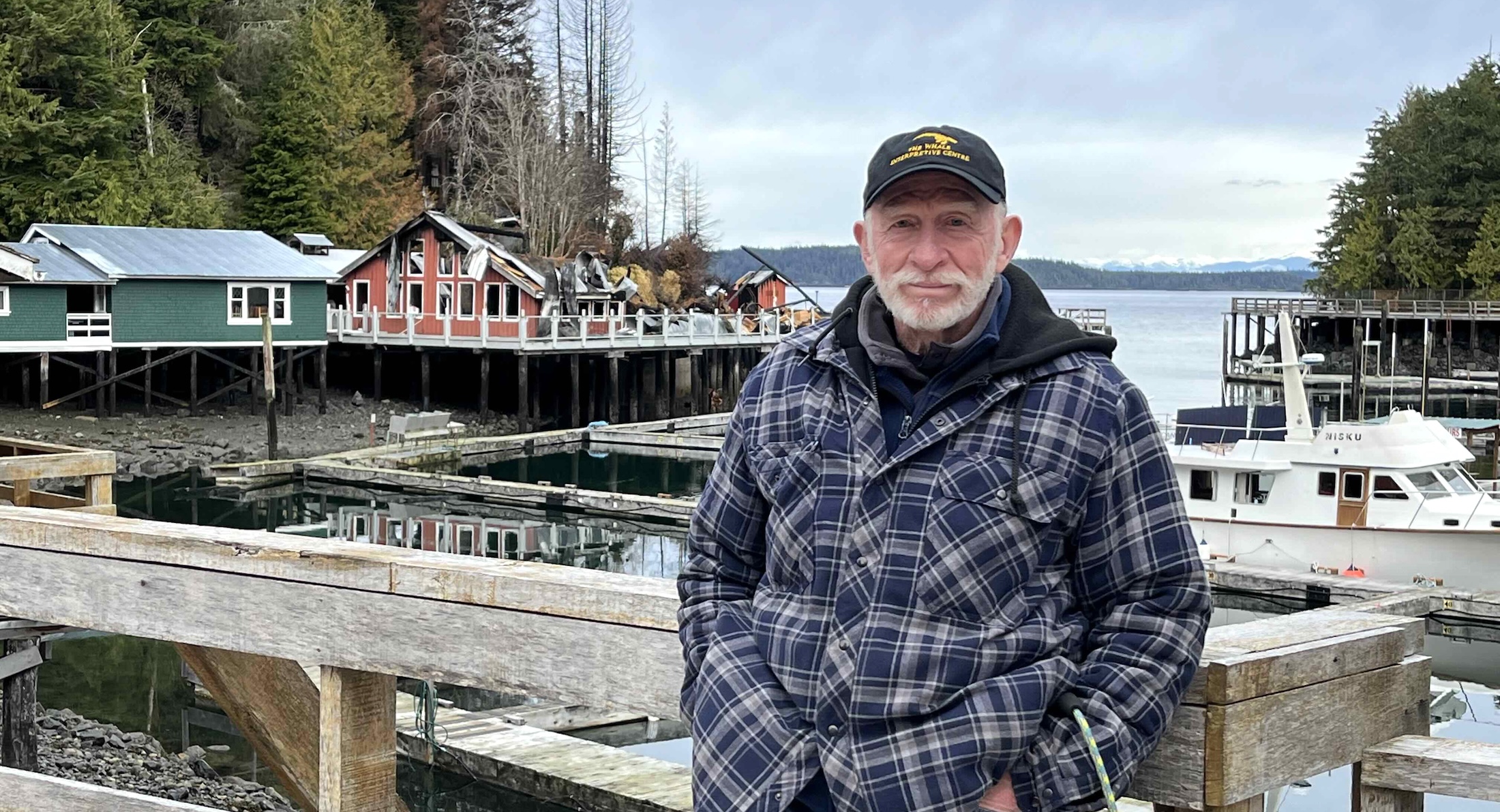 Jim Borrowman, a man in his mid-70s with light skin tone and a white beard, wears a ball cap, jeans and a plaid jacket. He stands on the boardwalk at Telegraph Cove, the burned-down Whale Interpretive Centre in the background.