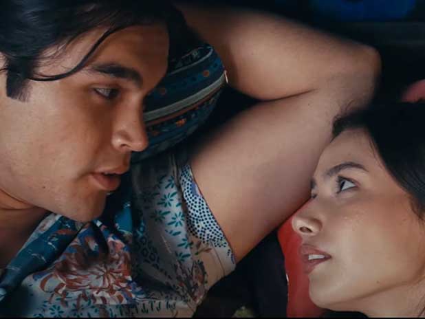 Actors Joshua Odjick, left, and Tatyana Rose Baptiste, right, are speaking closely together as they lie in colourful pillows. Odjick has medium skin and long black hair tied back. He is wearing a colourful shirt with a blue, white and red floral pattern. Baptiste has long dark hair, medium skin and is looking up at Odjick, mid-speech.
