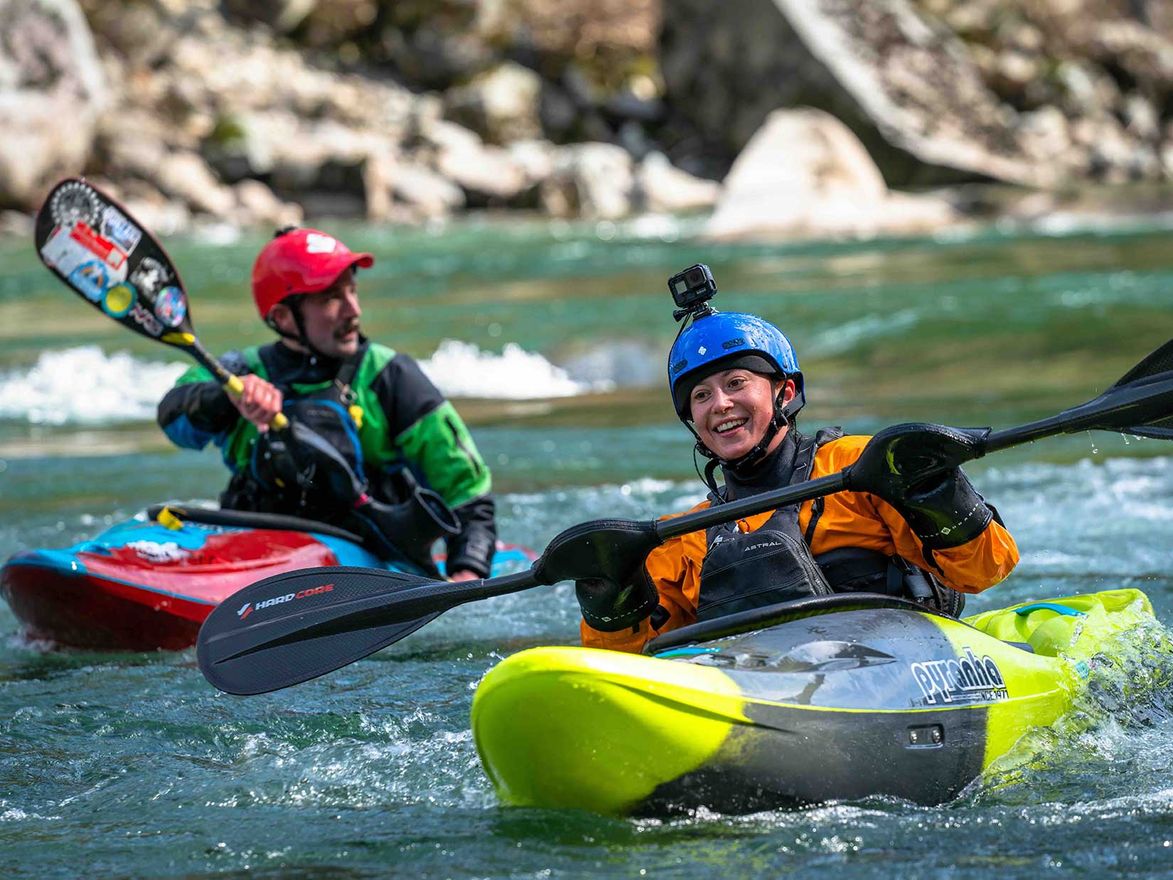A man and a woman are kayaking down a river. The woman, Alena, is first and in focus. She has a light skin tone and is in a yellow kayak, orange coat and blue helmet. She has a camera affixed to her helmet.
