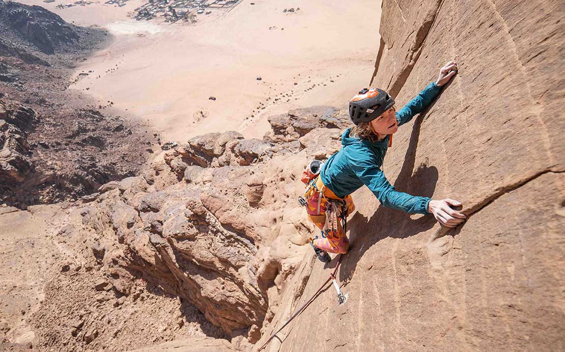 A person with light hair and skin tone looks up toward the camera while climbing a reddish-brown cliff face.
