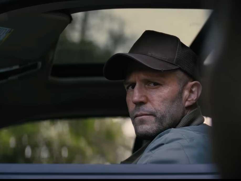 Actor Jason Statham wears a dark ball cap and a muted blue work jacket. He is looking out the driver’s side window of a large vehicle. He has pale skin and visible stubble.