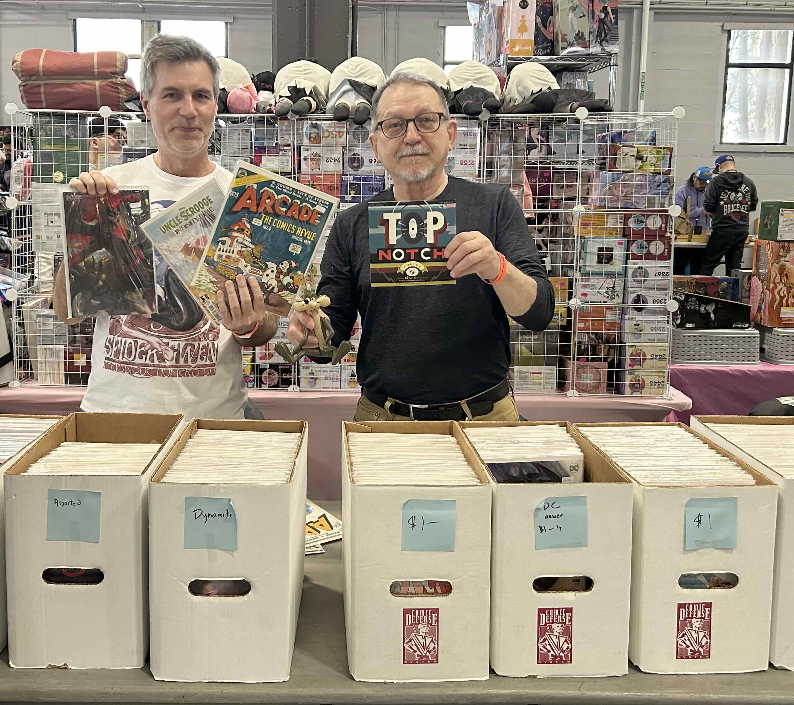 Shawn Conner, left, poses with his cousin Ken, right behind a grey table full of narrow white boxes containing comic books. Conner is wearing a white T-shirt with a red graphic and holds up three comic books. Ken is in glasses and a black top. He is holding a coyote figurine and a comic book in the other hand.