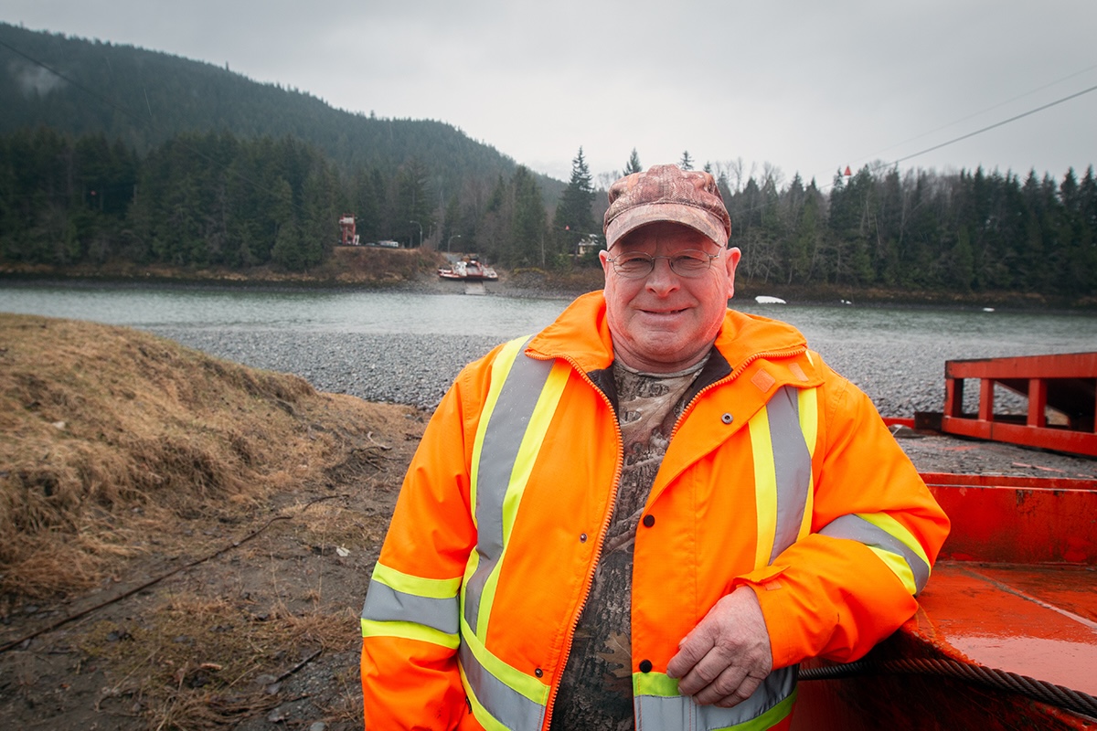 An older man wearing glasses, a camouflage hat and shirt, and an orange high-visibility jacket looks at the camera and smiles. He is leaning on a large, red piece of equipment and behind him there is a river.