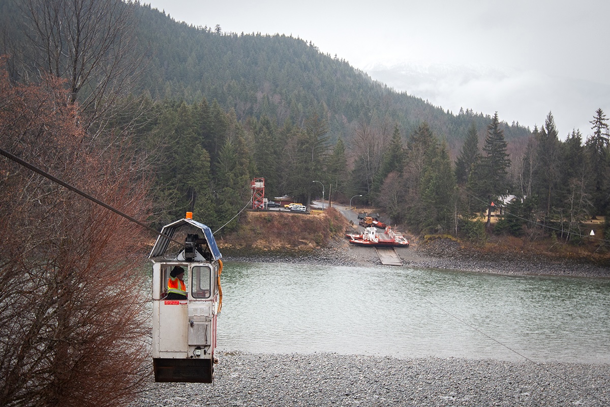 A small metal car hangs from a cable that is strung across a river. There are forested mountains in the background.