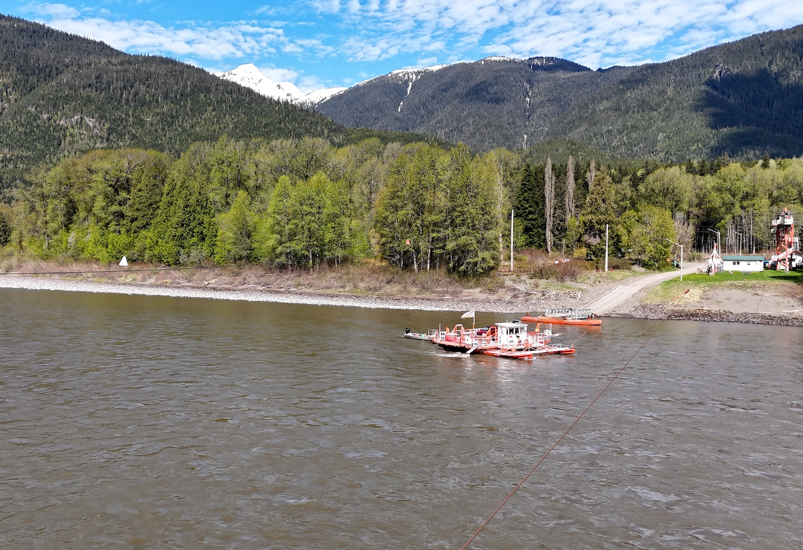 A photo taken from a high angle of a wide river with a white and orange vessel moving across it. Across the river there is forest and mountains beyond.