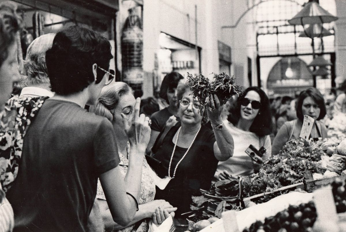 A black-and-white photo from 1978 is shot from inside a public market in Bologna, Italy. The market is crowded with women, and Marcella Hazan stands at the centre beside a produce stand, holding up a bunch of greens.