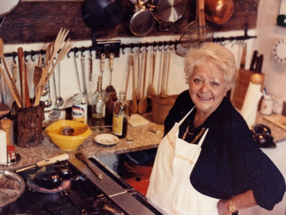 Marcella Hazan has short wavy white-blond hair, a medium skin tone and brown eyes. She smiles up the camera and is wearing a black shirt under a white apron. She is standing in a crowded kitchen before a gas stove.