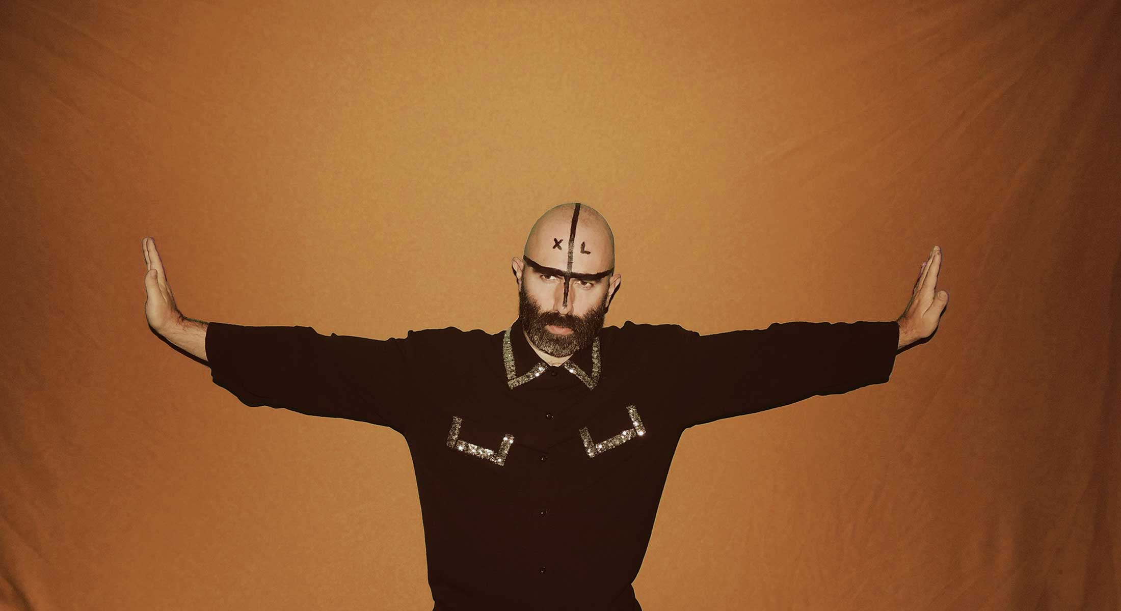 Jeff Cancade wears a black top with shiny flourishes lining the collar and two breast pockets. They are posing with their arms outstretched, looking to the middle distance away from the camera. They have a dark beard and are wearing black greasepaint across their face and head.
