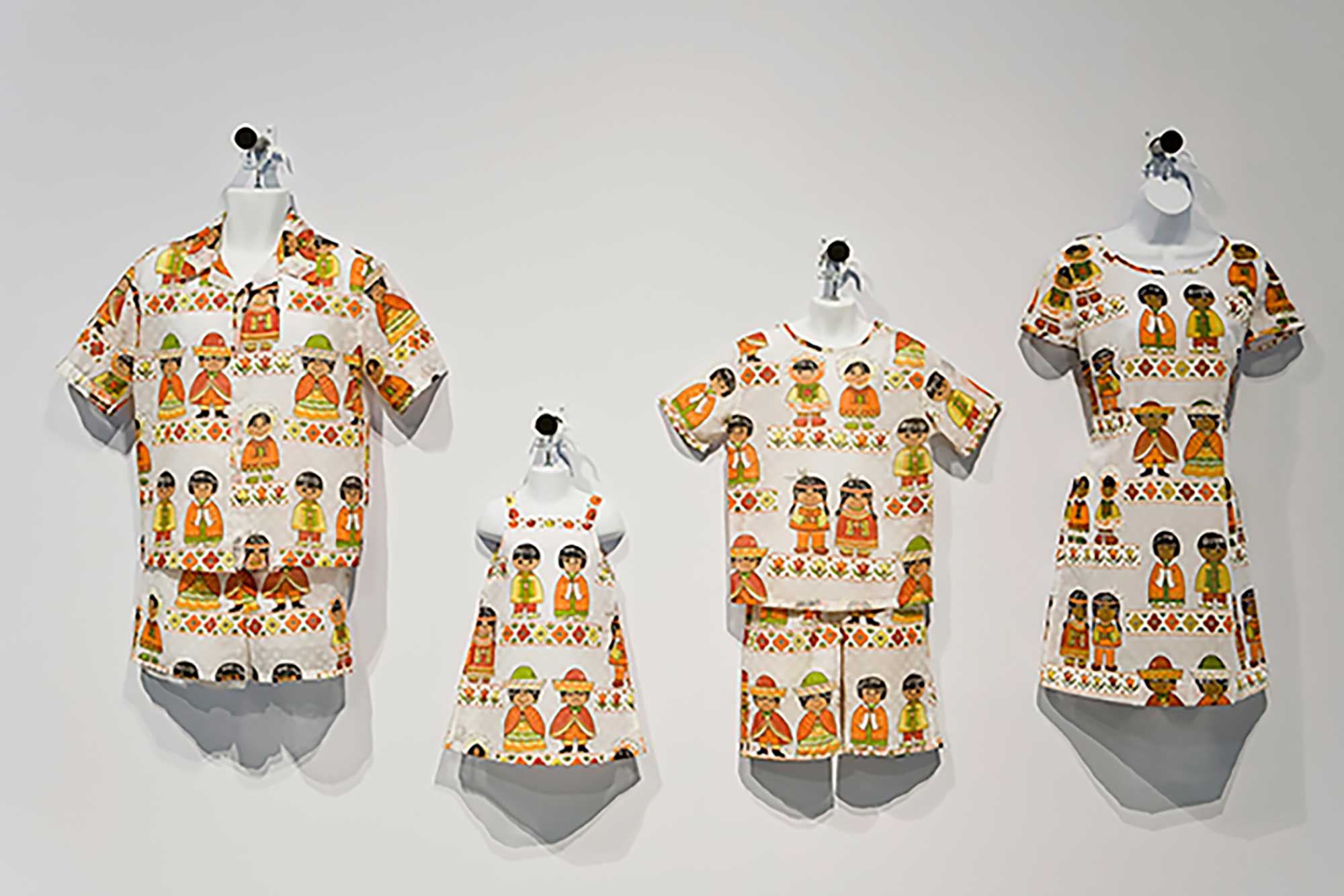 Four outfits that conjure the image, from left, of a father, a young daughter, a son and a mother hang on small white mannequins against a white wall. They are all attired in summertime outfits made with the same patterned fabric featuring yellow, orange, green and white colourways reminiscient of the 1970s featuring racist caricatures of people from around the world.