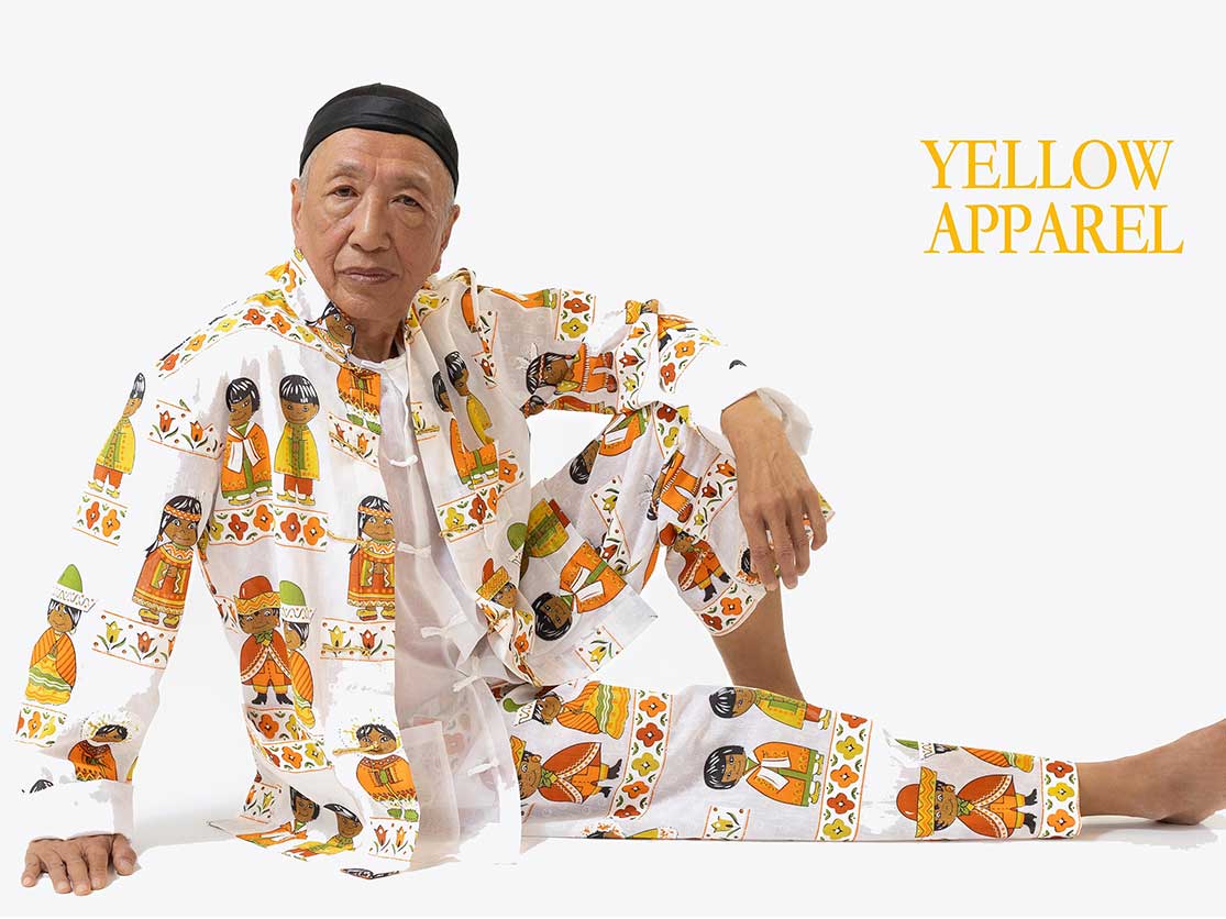 Paul Wong has a medium skin tone and is wearing a black head scarf. He is wearing a loose-fitting, traditional Chinese-style suit in a fabric featuring dated cartoon characters of people in the traditional Mexican, Inuit, Native American and Japanese clothing in yellow, orange, red and green colour schemes against a white background. 