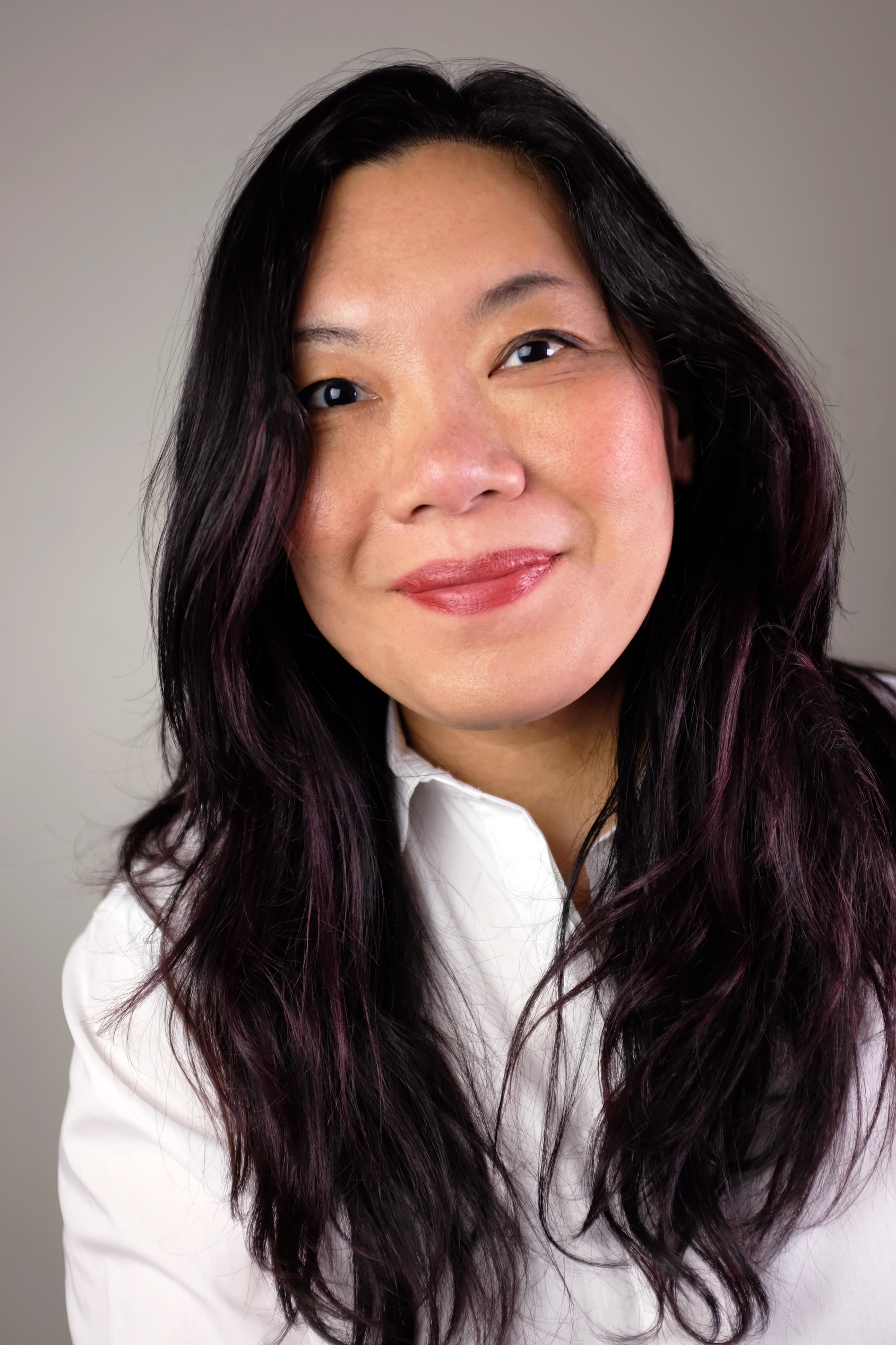 Donna Seto has long wavy black hair and a medium skin tone. She is smiling at the camera and seated against a grey studio background and wearing a white button-down shirt.