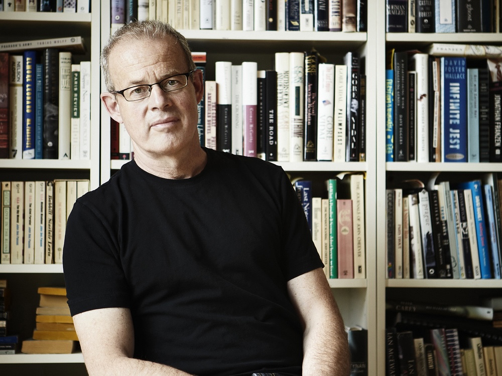 A portrait of a man with light skin tone and short, greying hair, wearing glasses and sitting in front of a shelf full of books.