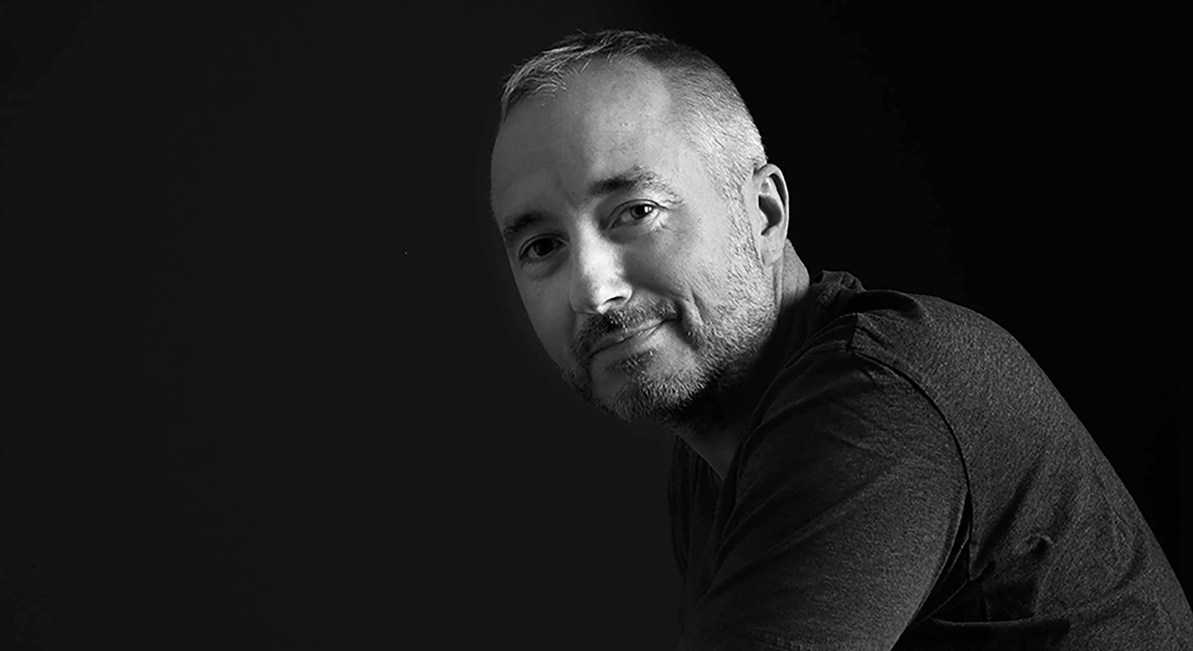 A black-and-white portrait of Pete Crighton features Crighton facing the left side of the frame in a dark T-shirt. He turns toward the camera, smiling gently. He has short greying hair and a beard.