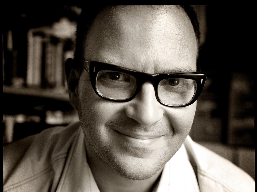 A warm-toned black and white close-up of Cory Doctorow, a man with short dark hair wearing dark thick-framed glasses.