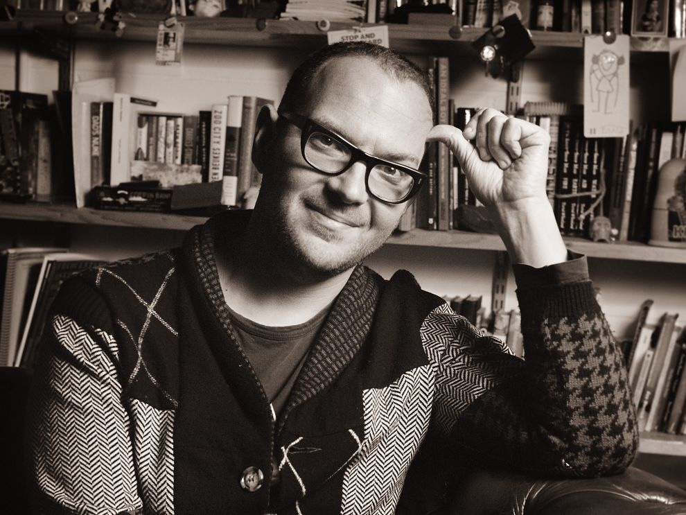 A black and white photo of Cory Doctorow, a man with light skin tone, close-cropped hair and black-framed glasses, sitting in a library, wearing a sort of knit patchwork blazer cardigan.