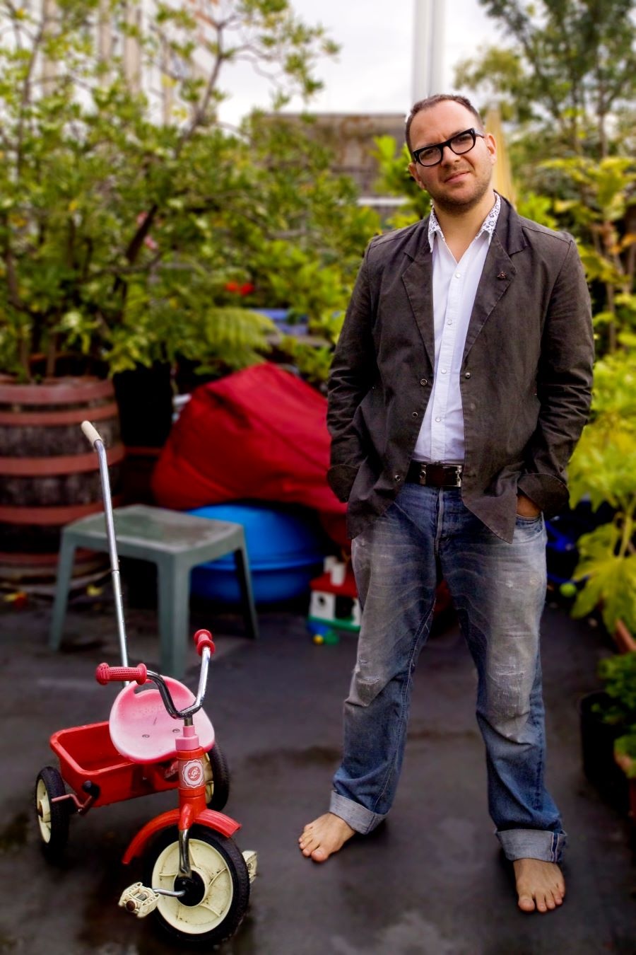 Cory Doctorow, a man with light skin tone, close-cropped hair and black-framed glasses, stands barefoot in jeans next to a child&rsquo;s red tricycle.