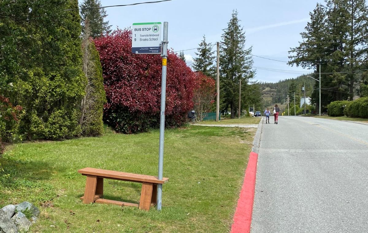 A brown bench is affixed to a bus stop pole in the grass. On the right the curb is painted red next to a wide road where pedestrians are walking in the distance. 