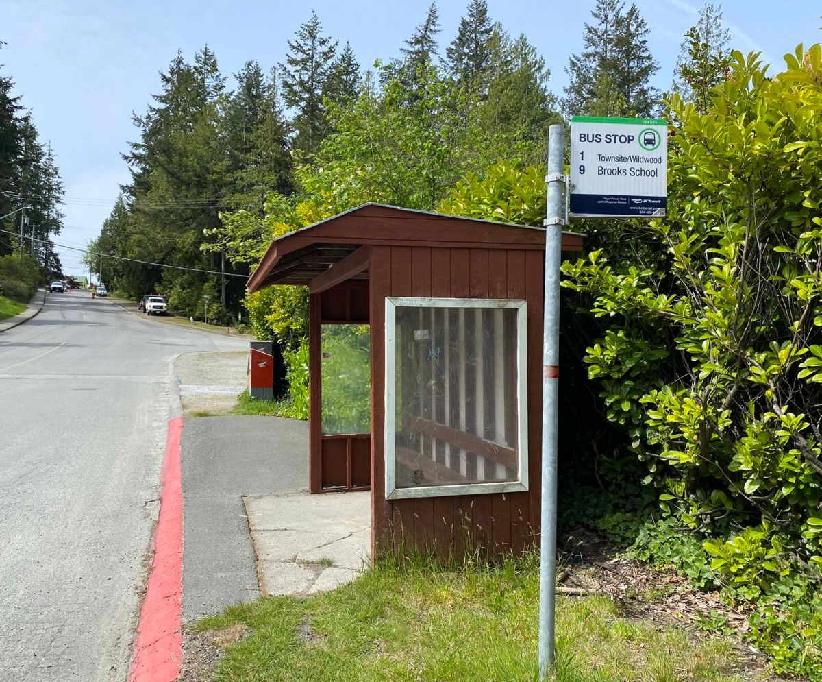 A brown shed with a glass windows stands by the side of the road next to a silver pole that indicates the bus stop. In the background is a fork in the road and a Canada Post mailbox.