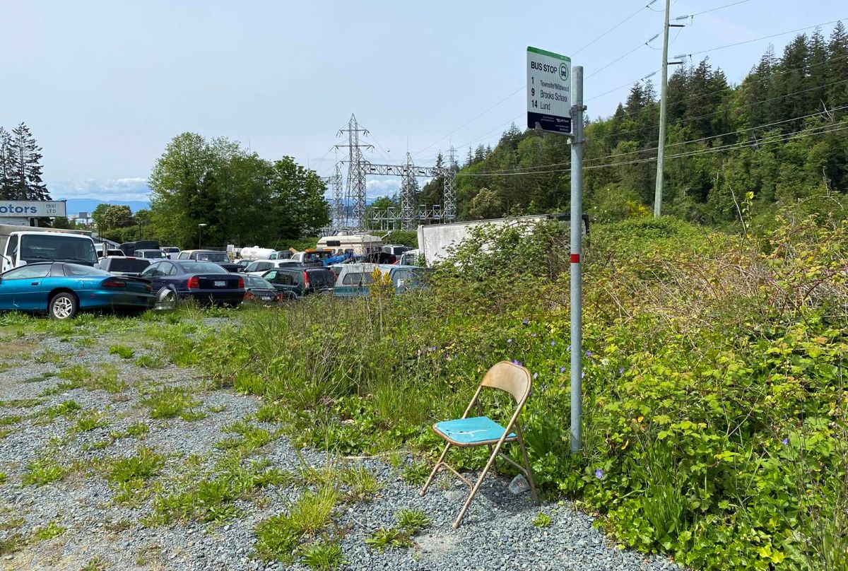 A folding plastic chair with gold trim and a turquoise seat stands in the gravel next to a bus stop amidst shrubbery. In the background are an assortment of cars parked in a lot.