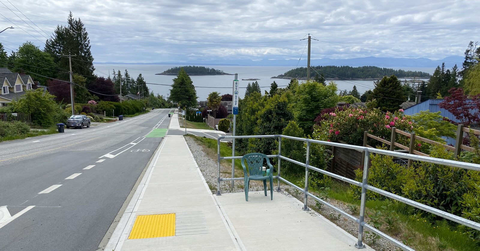 A green plastic chair stands next to a bus stop pole on a sidewalk next to a roadway. Behind it is a view of the ocean and nearby islands.