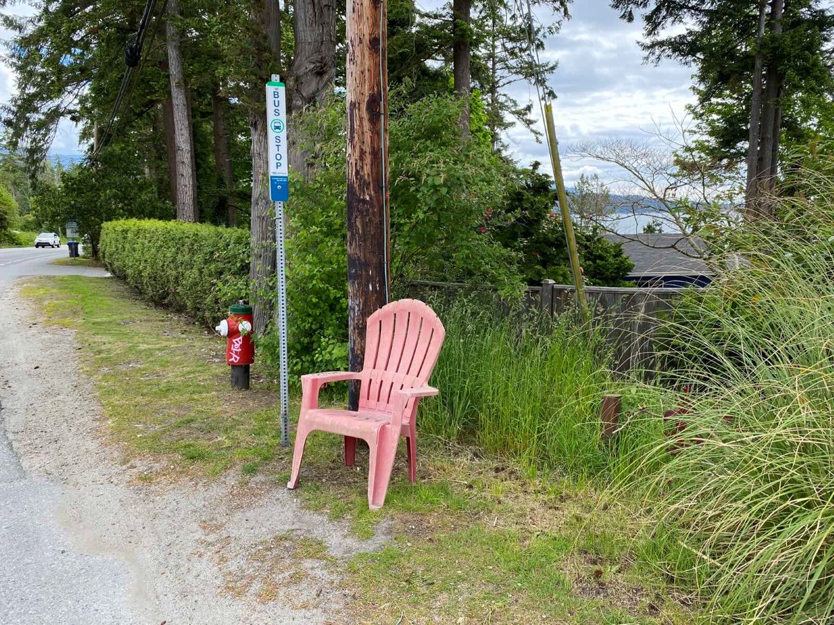 A faded red plastic lawn chair sits against a wooden electric pole in the grass by the side of the road next to a bus stop pole. The water is visible in the background.