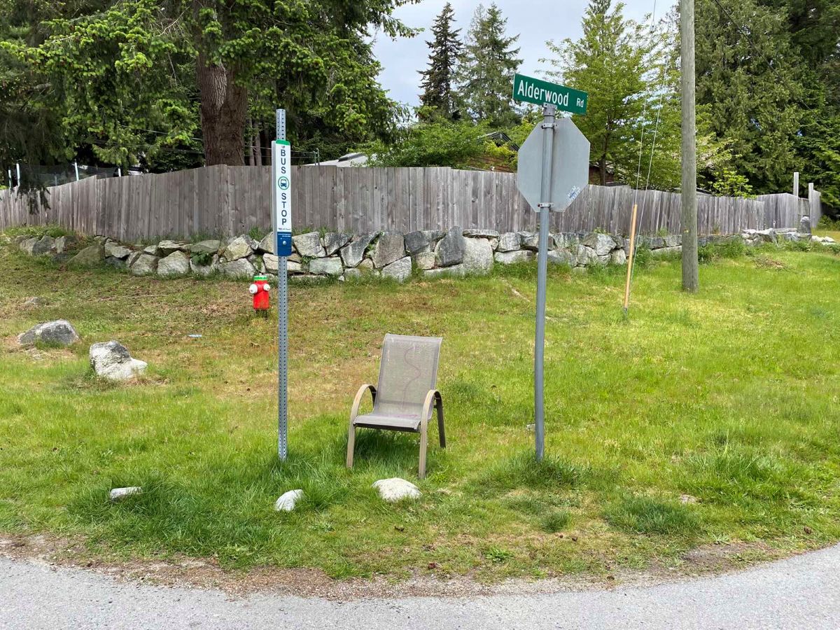 A taupe chair stands next to a bus stop pole in the grass beside a stop sign.