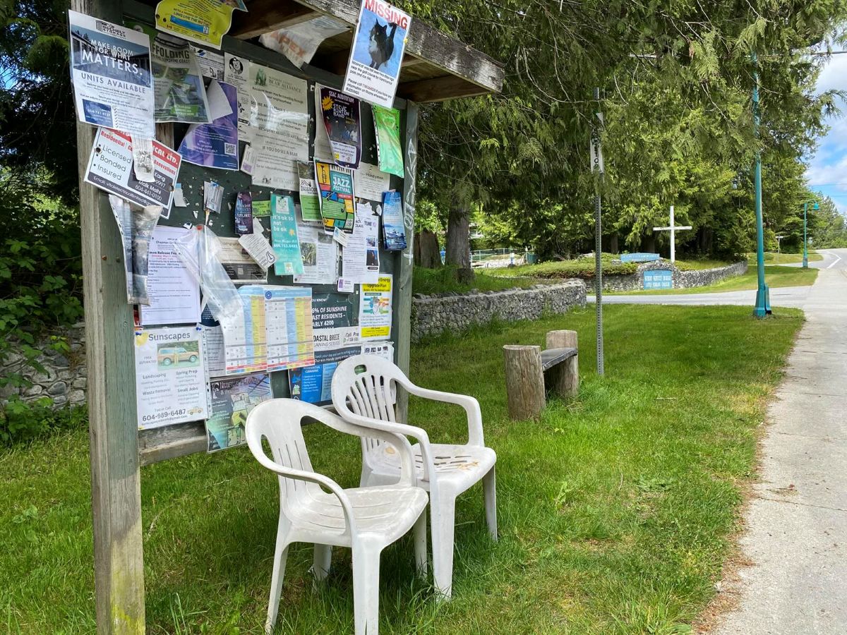 Two white lawn chairs are positioned in front of a community message board. To their right, there is a wooden bench and a bus stop sign on a metal pole.
