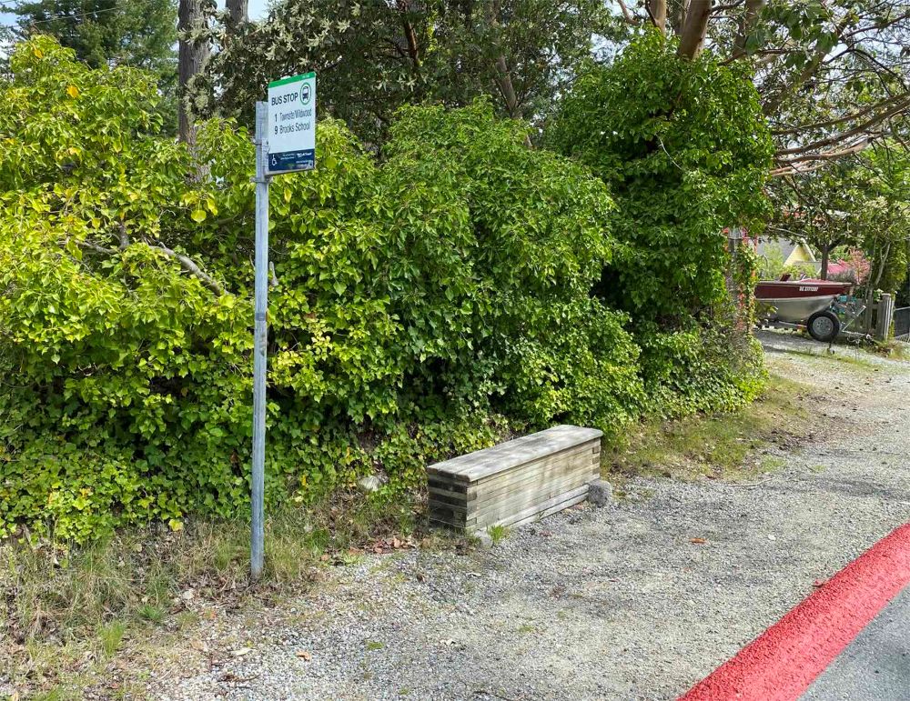 A small solid wooden bench sits near a bus stop road near a gravel pathway.
