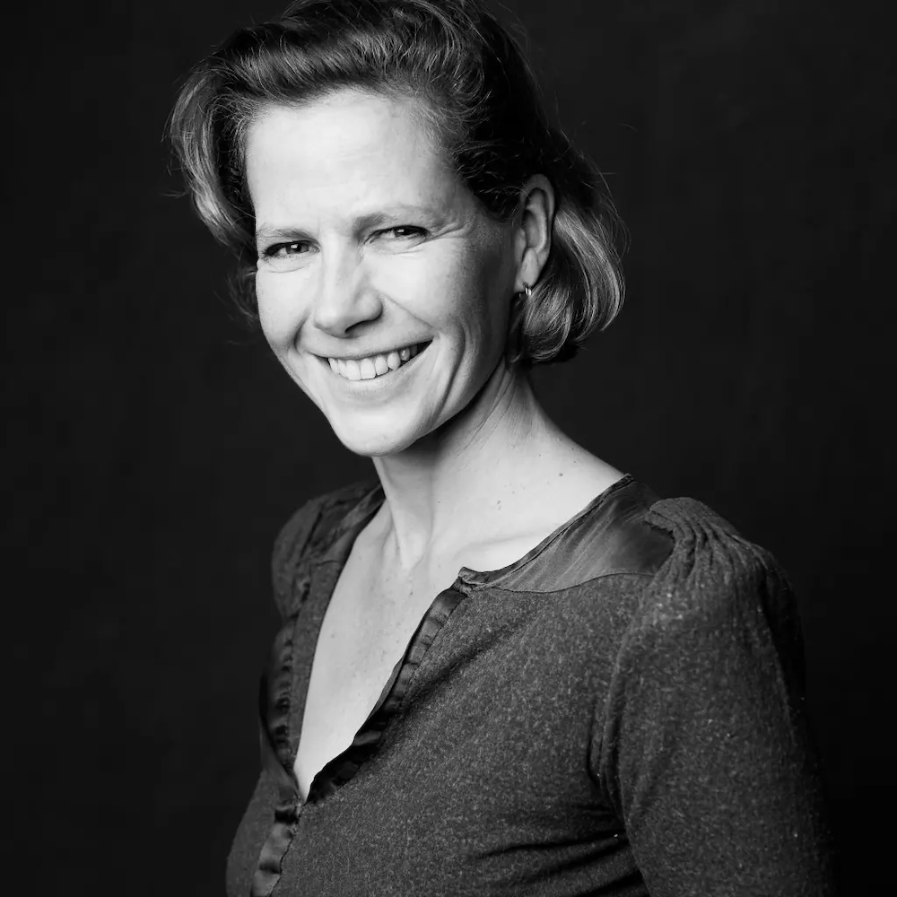 A black and white portrait of Laura Spinney, a woman with light skin tone, short wavy hair and a V-neck top. She is smiling at the camera.