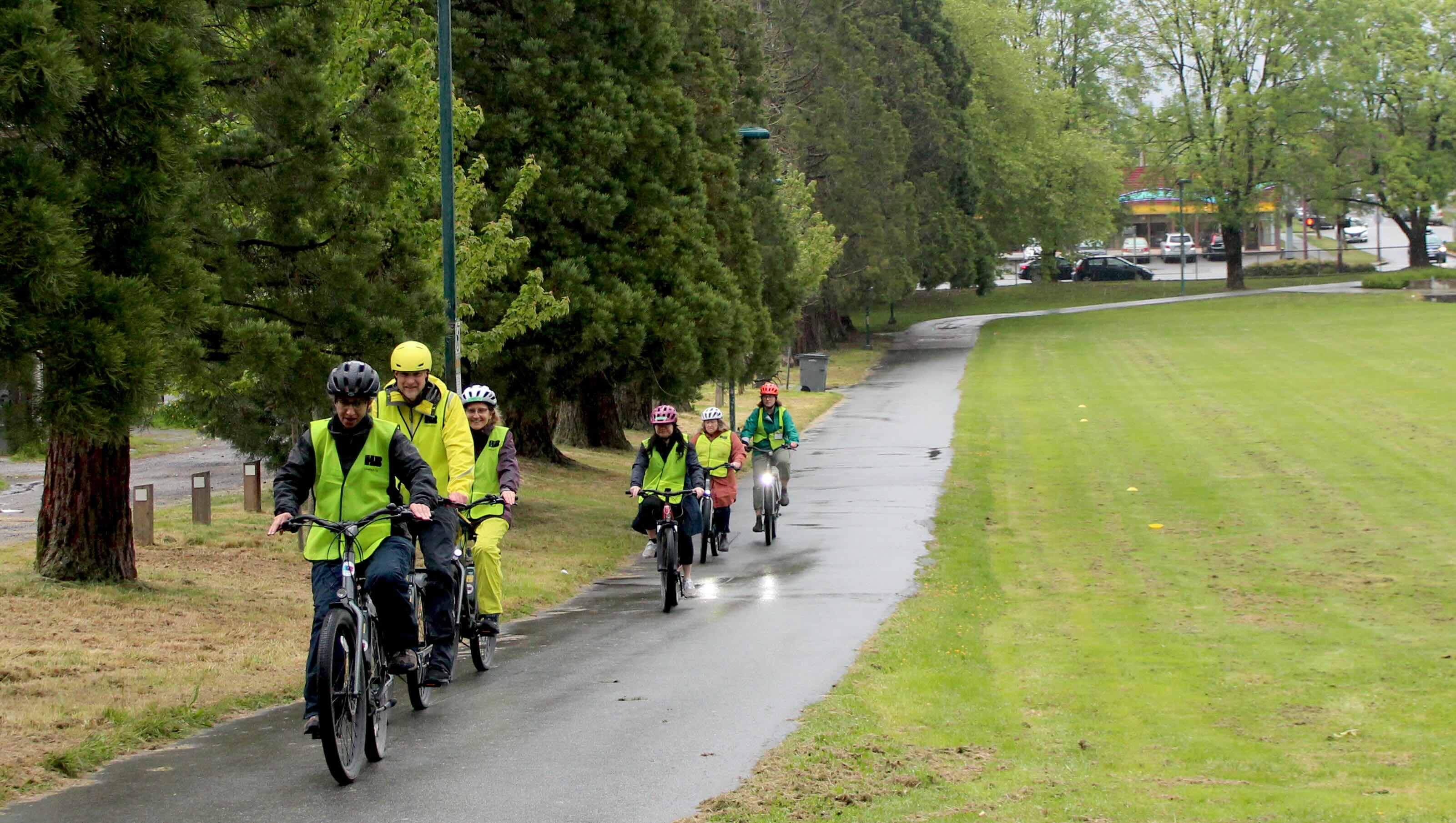 A group of six cyclists on e-bikes are riding in single file along a cement pathway through a park. They are wearing bike helmets and large neon yellow reflective vests.