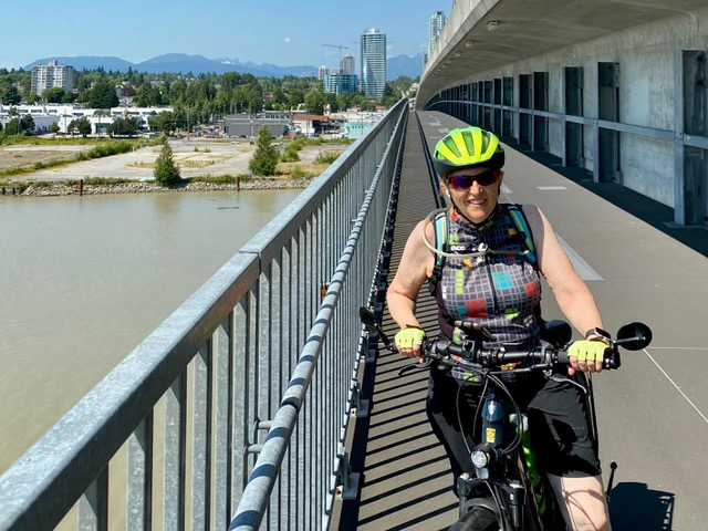 Karole Sutherland poses with her bike while riding across a pathway over water on her left. She is wearing a neon yellow helmet and sunglasses.