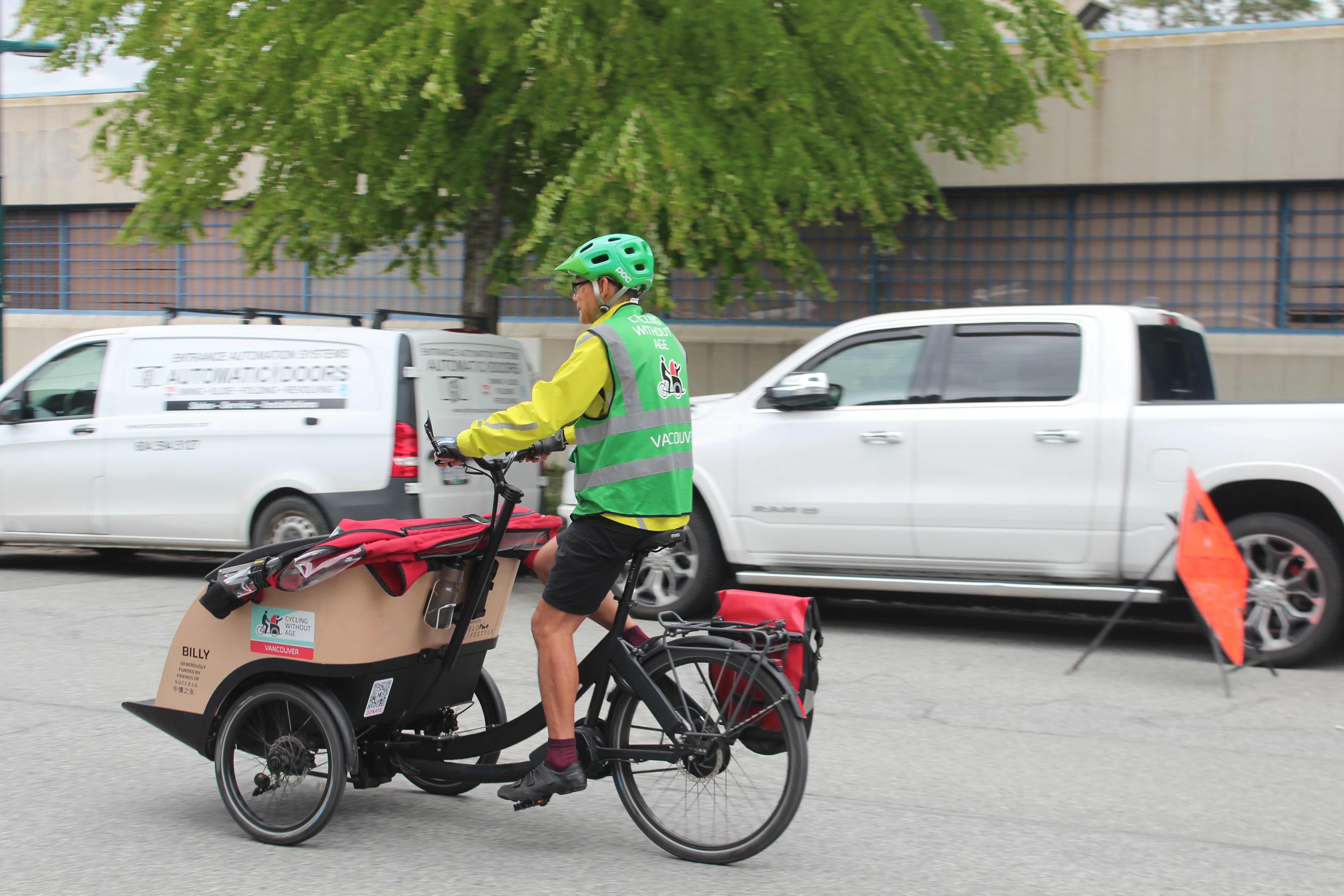 An older cyclist in a green helmet, green vest, yellow jacket and black shorts rides an electric bike with two wheels at the front on which a cargo area is mounted for two passengers to sit.