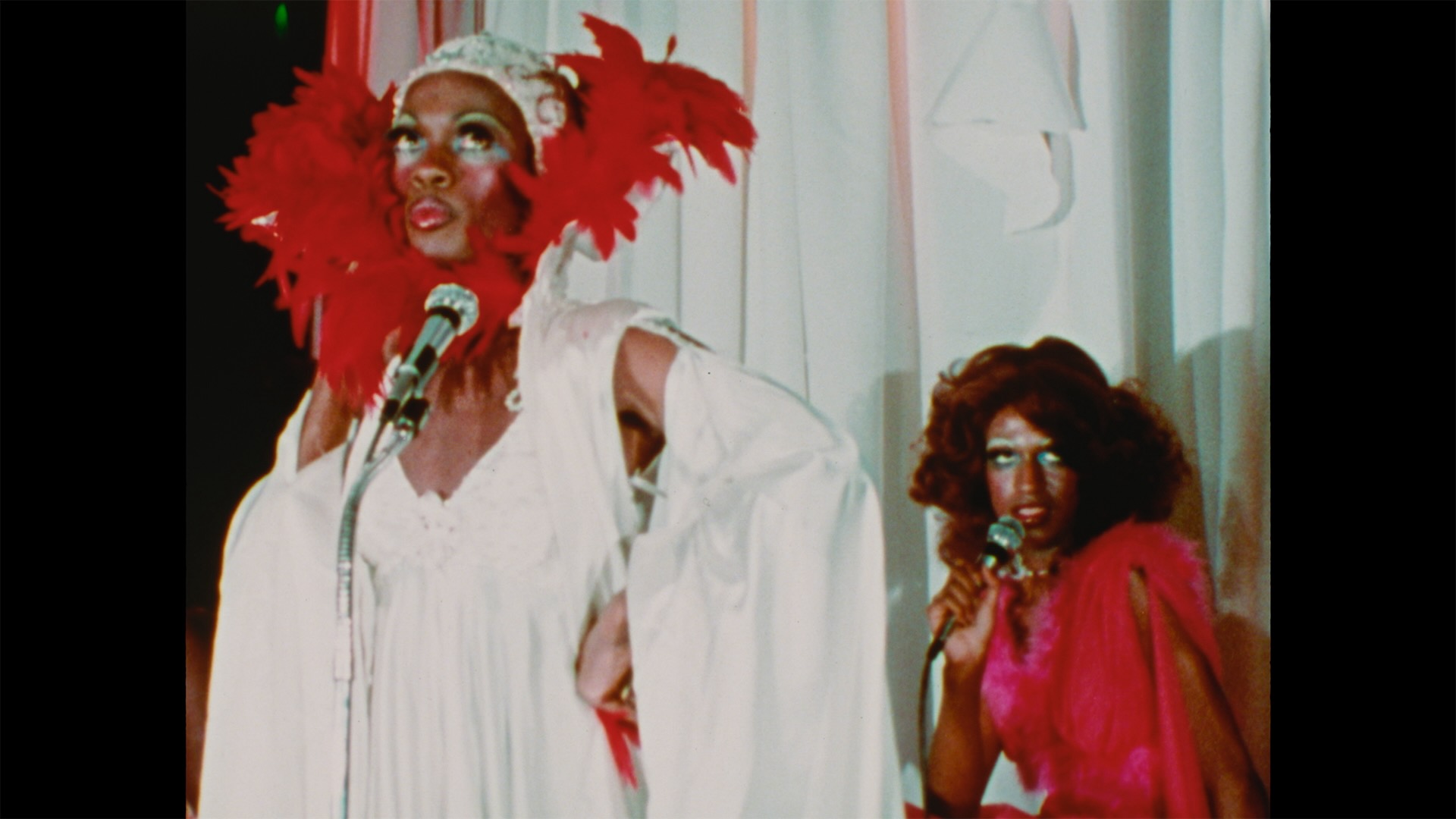 Michelle Ross, left, is a drag performer wearing a white headpiece, a white gown and a feather boa around her face. She has dark skin and is wearing light green eye makeup and exaggerated blush and lipstick. She stands onstage at a microphone and another performer, in red, is seated at a microphone behind her.