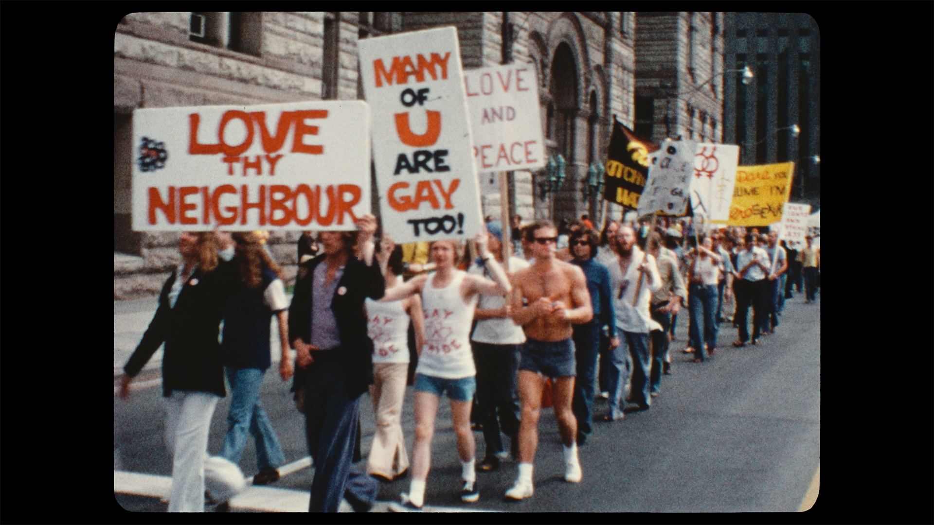 An archival still from 1973 depicts people in casual summer clothing marching down a Toronto street holding large white signs that read “Love thy neighbour,” Many of u are gay too!.” “Love and Peace” and more.