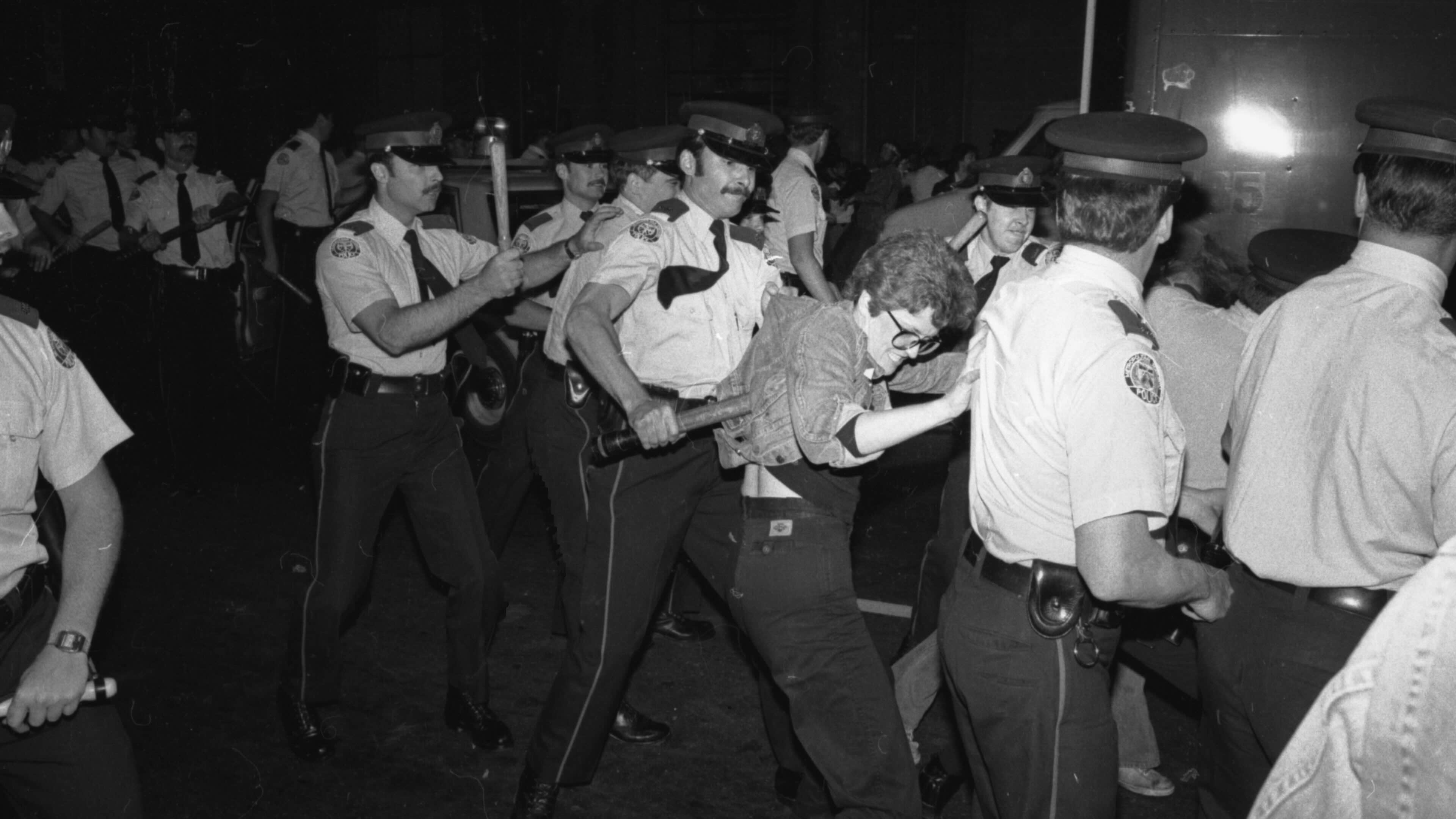 A black-and-white photo depicts a large crowd of police officers in white short-sleeved shirts, dark ties and police caps holding billy clubs. One is moving his club onto the back of a man shorter than him; he has short wavy hair and glasses.