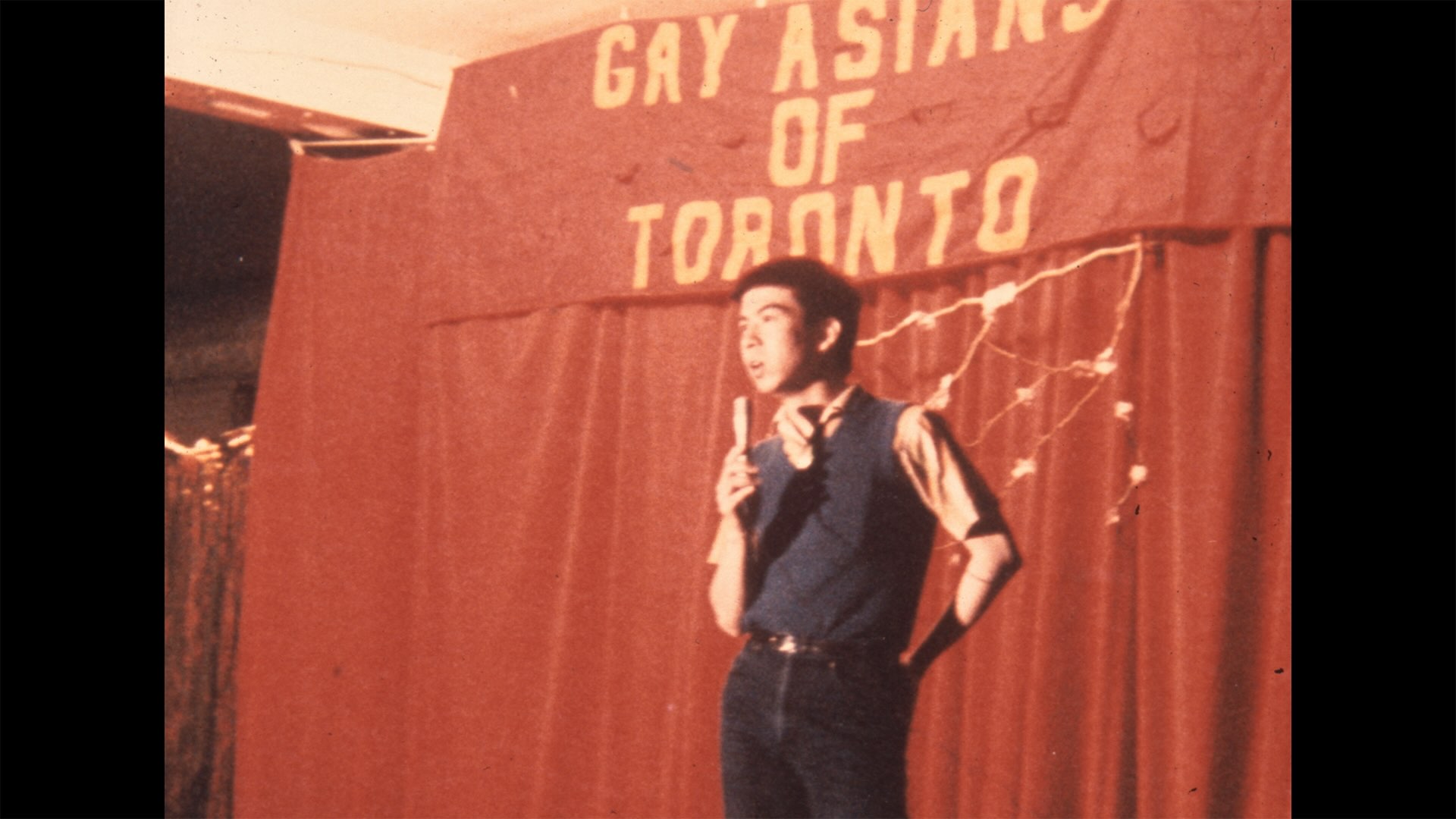 James Ma is a young man with short black hair and medium skin. He wears a navy sweater vest over a short-sleeved shirt and jeans. He is standing onstage, holding a microphone against a red curtain backdrop and a banner that reads “Gay Asians of Toronto.”