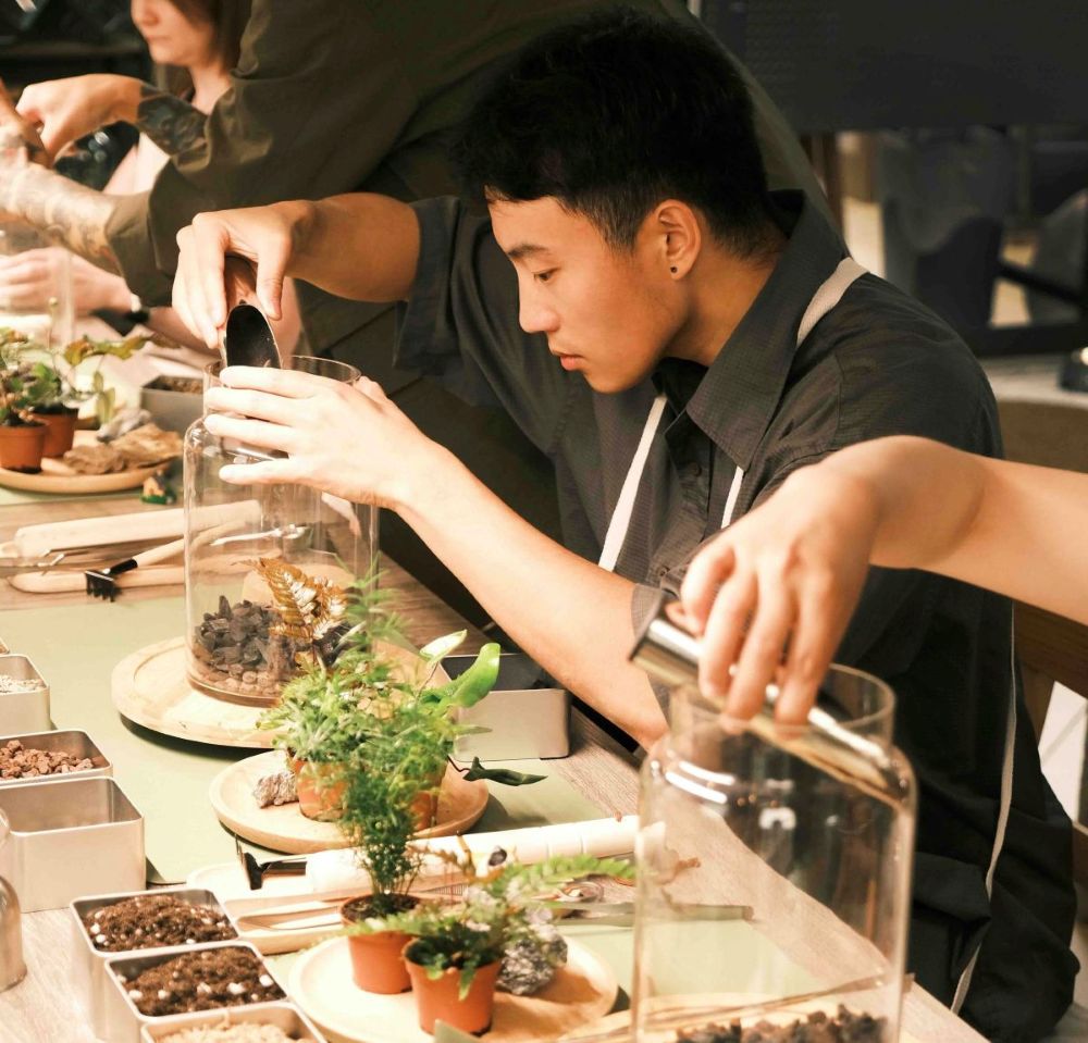 A young man with short black hair and a grey shirt is seated at a table using tools to arrange rocks and plants in a tall, wide glass vessel. He is surrounded by other people doing the same thing.