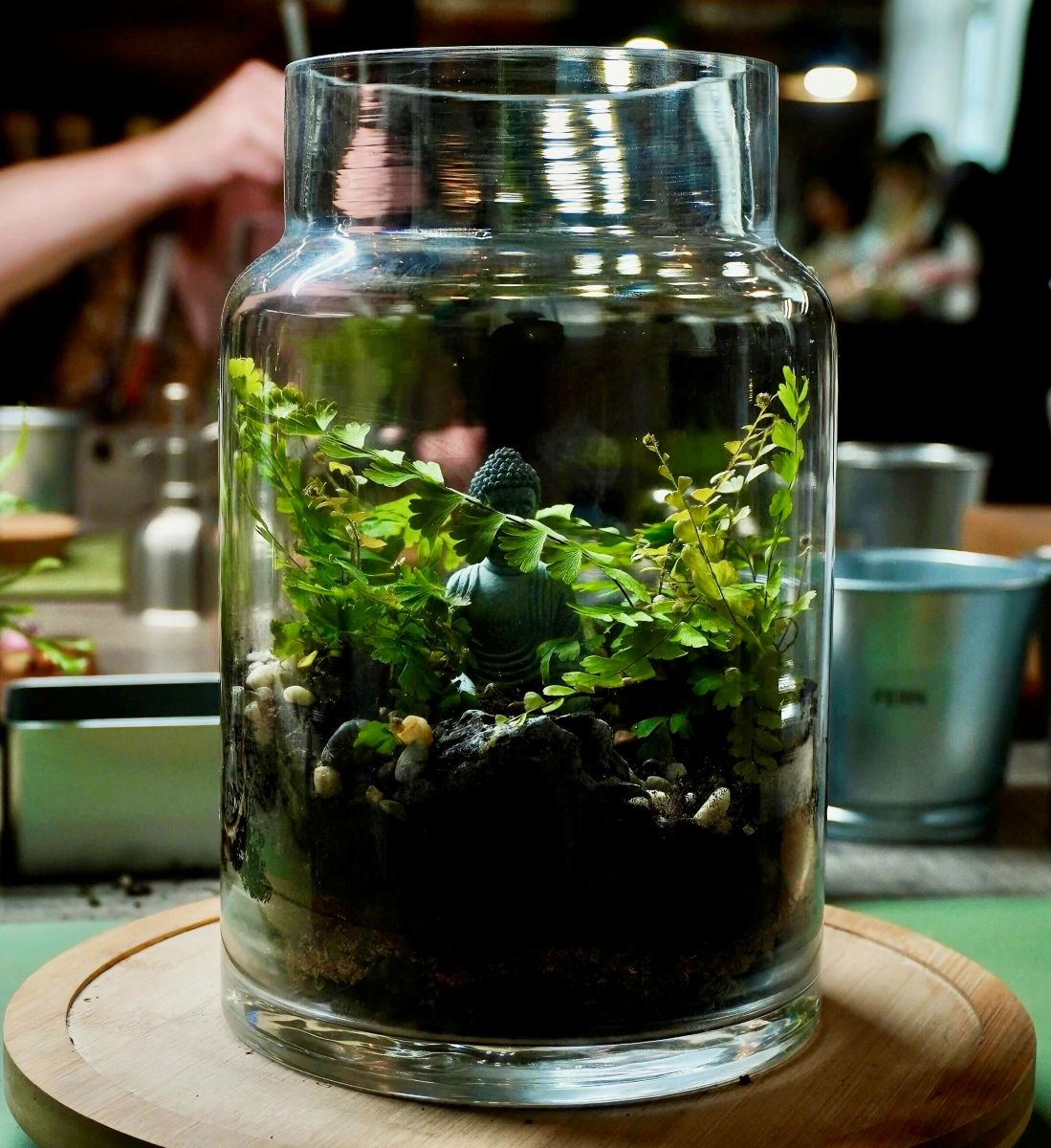 A photograph of a terrarium in a tall wide glass vessel featuring small, long leafy branches around a miniature grey statue of a goddess.