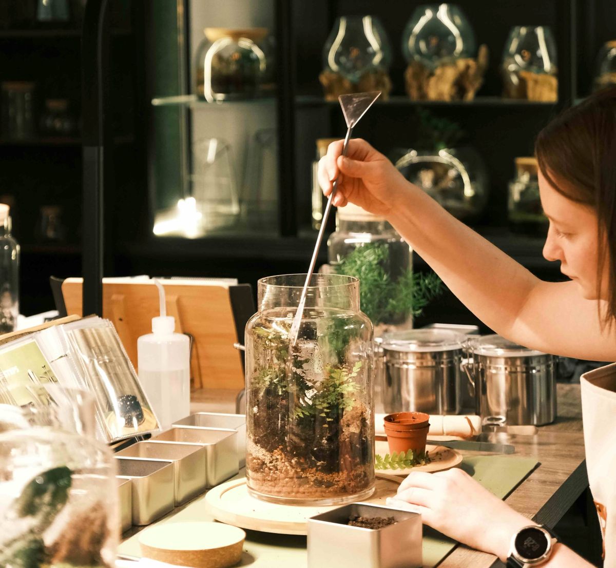 A woman with short brown hair is seated to the left of a terrarium-making table in an indoor space. She is using a long steel tool to arrange the plants inside a tall, wide glass vessel.