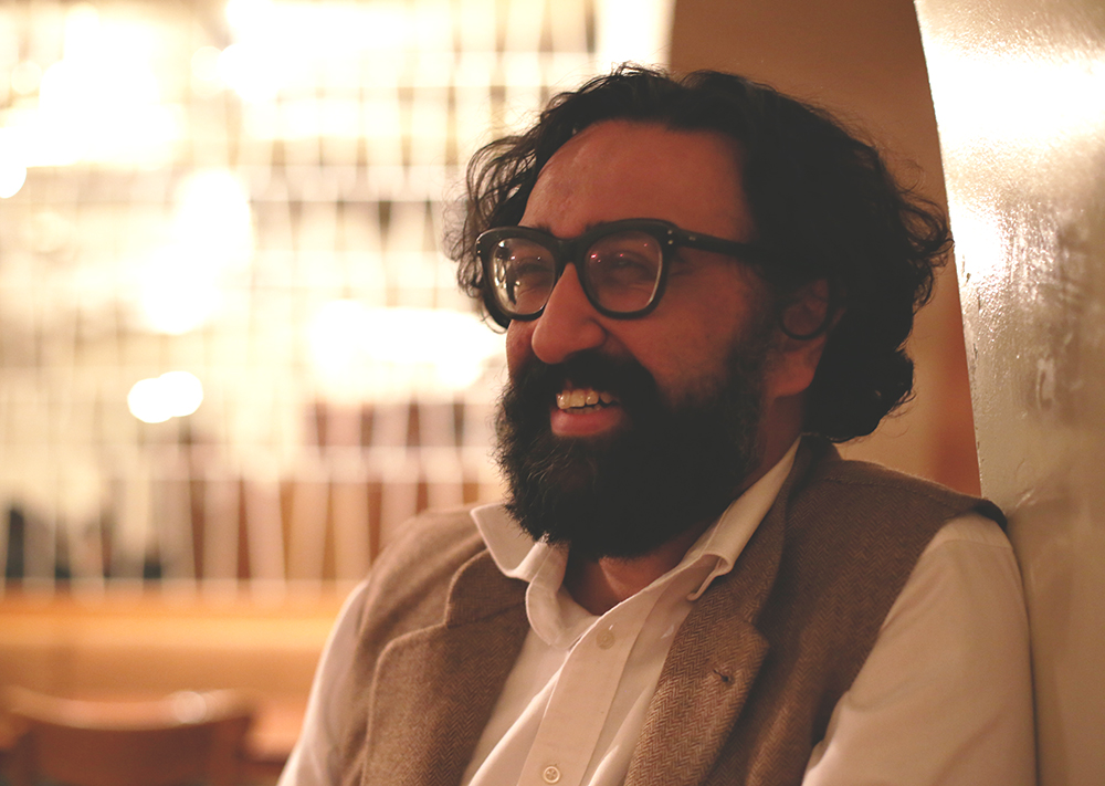Am Johal has a medium skin tone, short wavy hair, a beard and thick-framed glasses. He is seated in an indoor space, smiling. He is wearing a white shirt under a light brown vest.