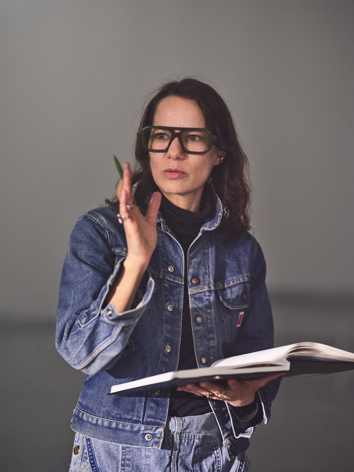 Justine Chambers has a medium skin tone and is wearing thick-framed glasses. She has shoulder-length brown hair and is wearing a denim jacket over a black top. She is holding a notebook in one hand and gesturing with the other. She is standing against a grey background.