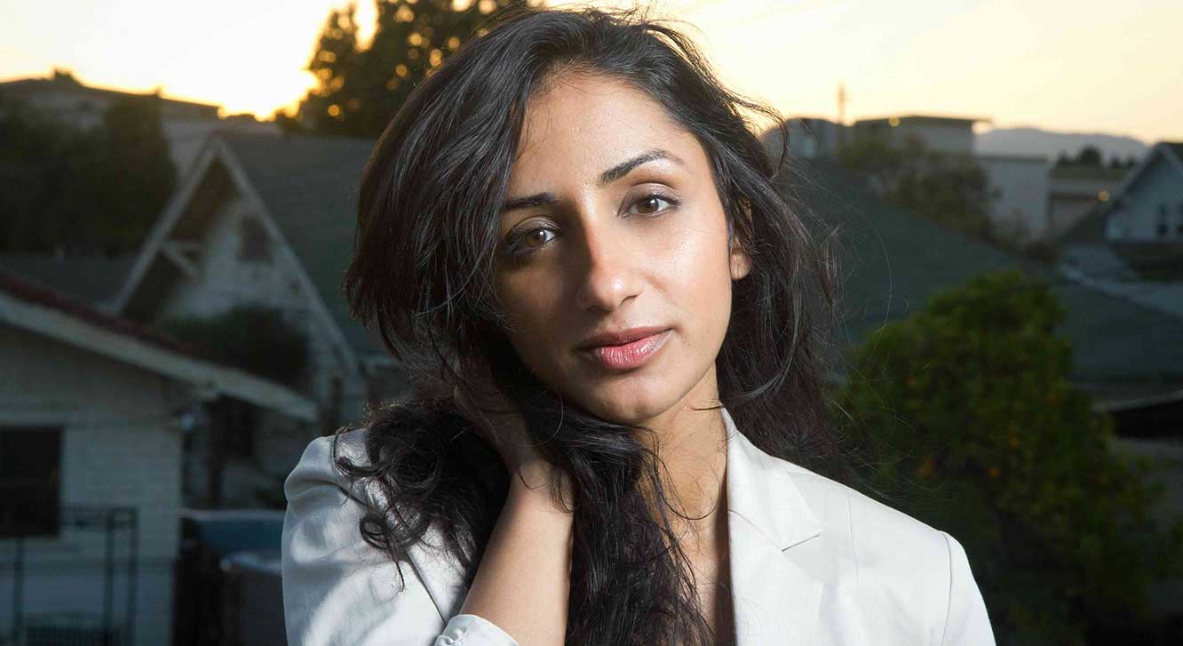 Kiran Deol has long, dark wavy hair and a medium skin tone. She holds one hand in her hair and looks at the camera. In the background is a greenscreen featuring photographs of a residential neighbourhood at sunset.