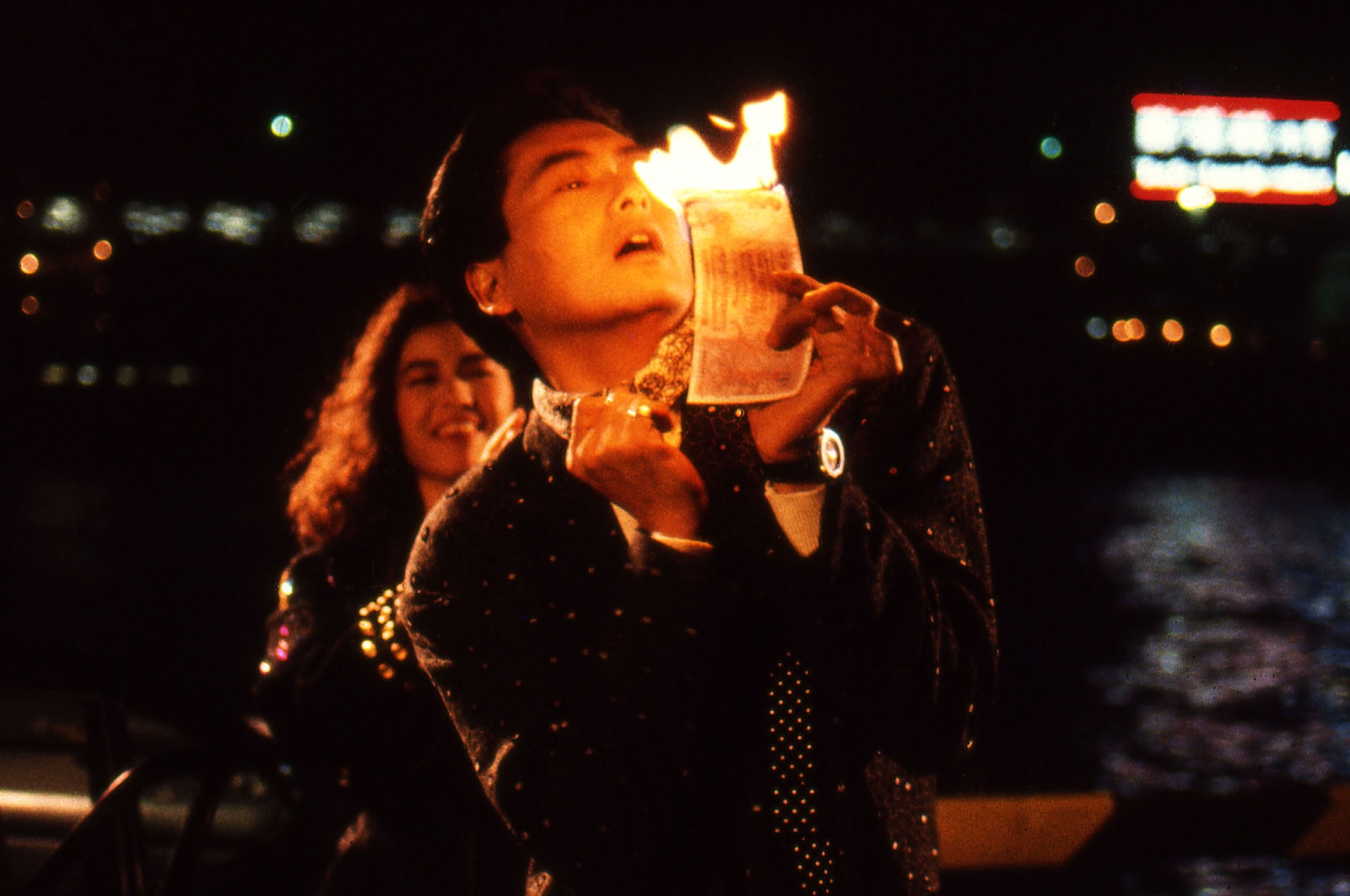 Actor Chow Yun Fat holds what appears to be a bill on fire while standing outdoors in a dense urban setting at night. Behind him, actor Cherie Chung is smiling.