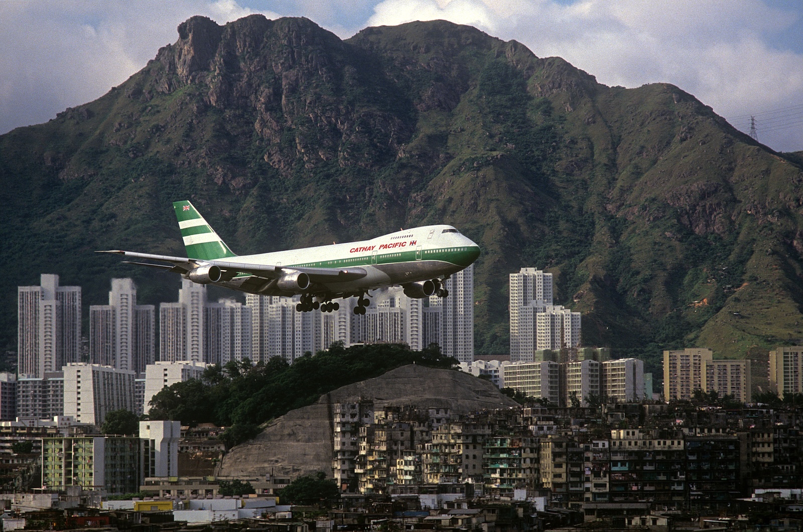 A large white and green Cathay Pacific plane flies against a bank of skyscrapers against a mountainous backdrop in Hong Kong.