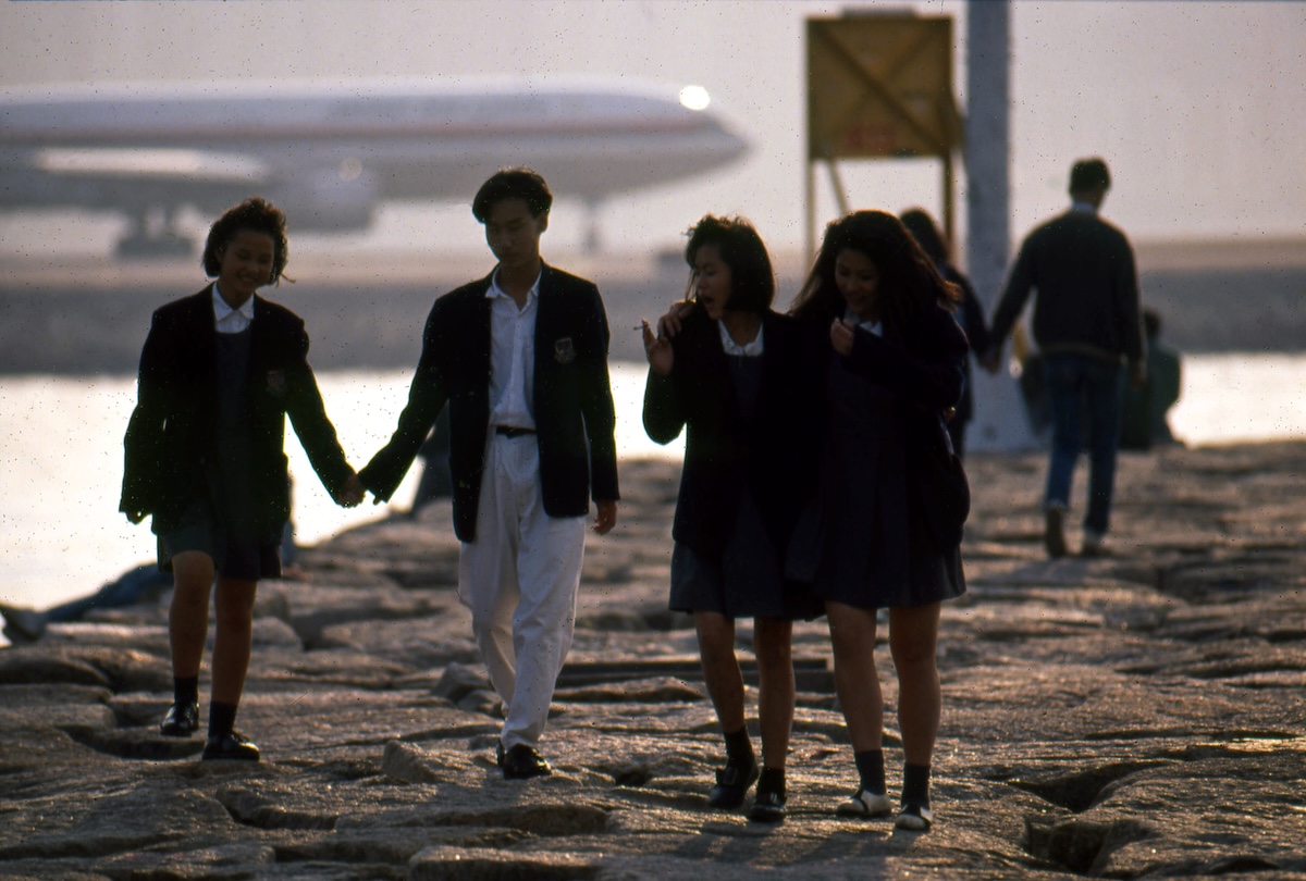 Four young people walk together on a grey concrete walkway set with oblong stones near the water in Hong Kong. The sky is grey and hazy.