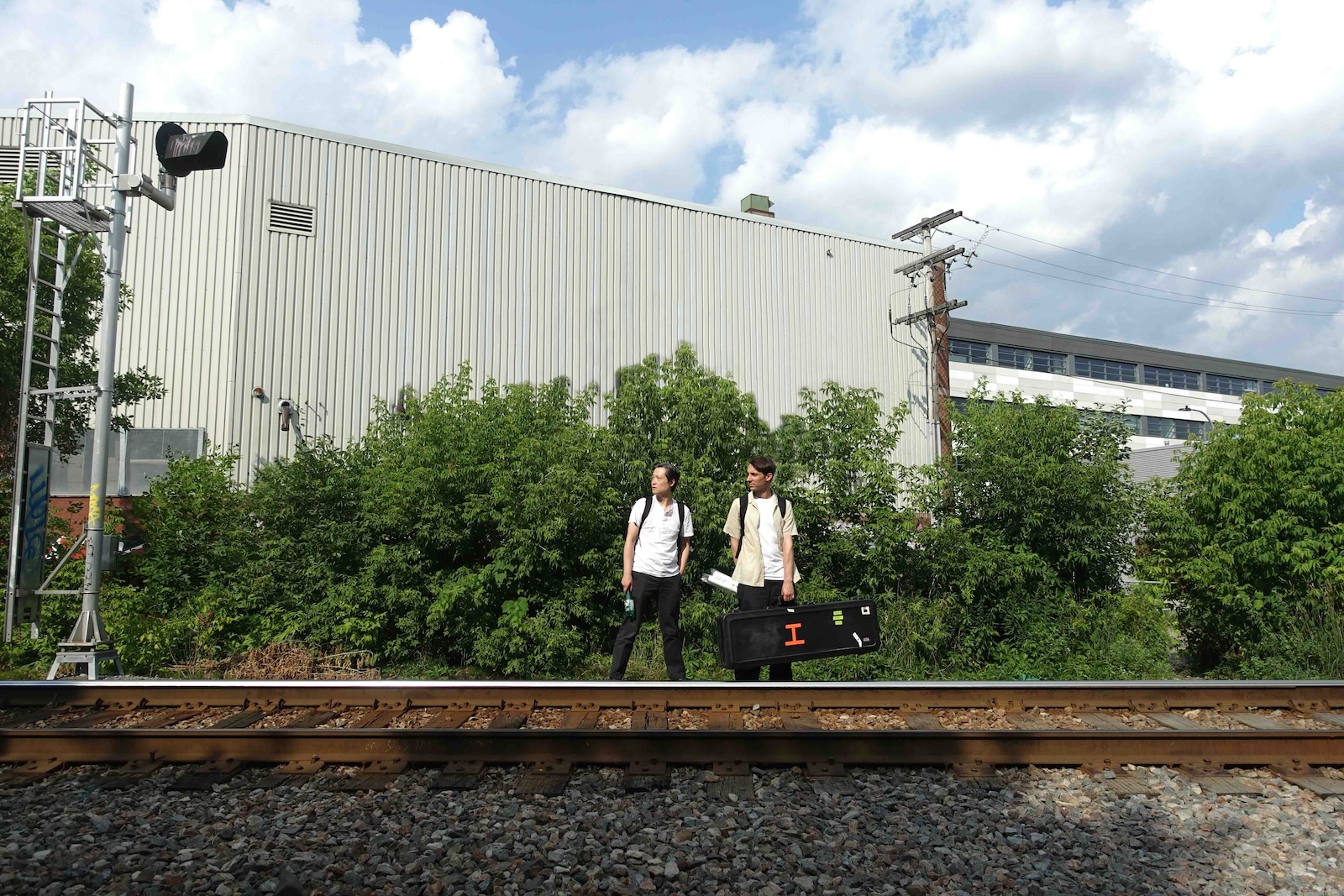 Tom Ng, left, and Joshua Frank, right, are wearing white T-shirts and black jeans. Ng has black hair and a light skin tone; he is wearing a black backpack. Frank has brown hair and a medium skin tone; he is wearing a light yellow short-sleeved shirt over his T-shirt and carrying a black guitar case. They are standing in front of a stand of shrubs behind a railroad track.
