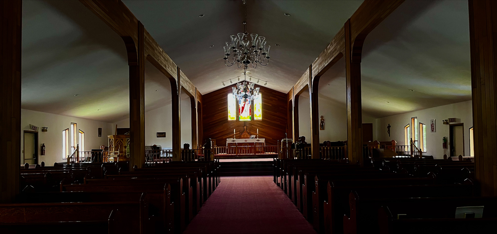 The interior of a church. The roof is simple and peaked. There is one central aisle, flanked by paired wooden columns with gentle, shallow arches. Behind the altar, the wall is decorated with cedar planking set at the same angle as the roof. 