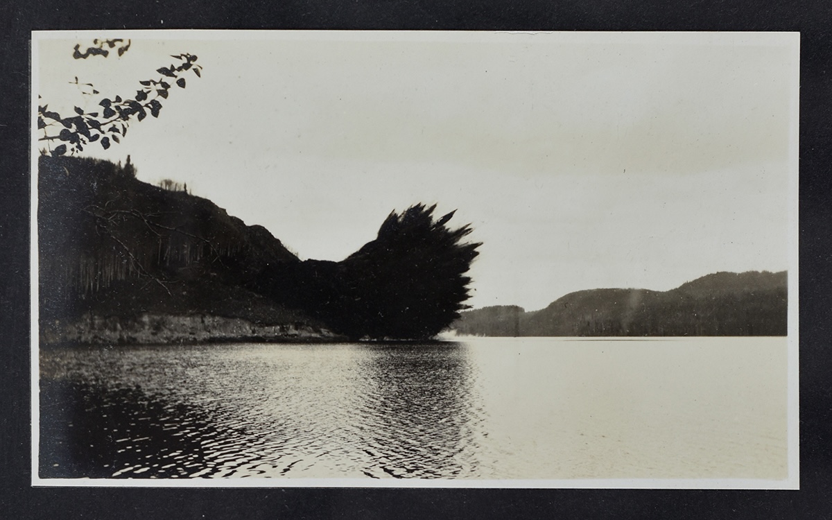 A black and white photo shows an expanse of water in the foreground and a hillside in the background. An explosion appears to be emanating from the hillside.